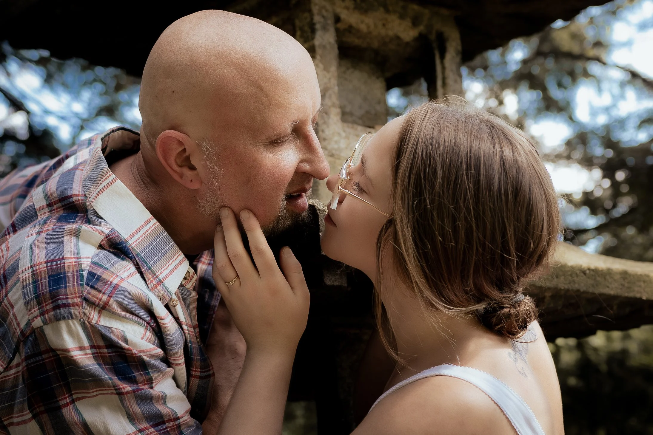 A man and a girl are touching foreheads and smiling in an outdoor setting with trees in the background.