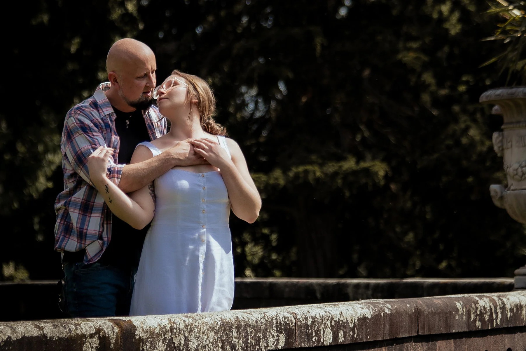A man with a shaved head and beard embracing a woman with glasses and long hair, both standing outdoors during daytime. The woman is wearing a white dress, and the man is wearing a plaid shirt over a black T-shirt. They are close to each other, looking into each other's eyes, with a dark, blurry background of trees and a stone railing.