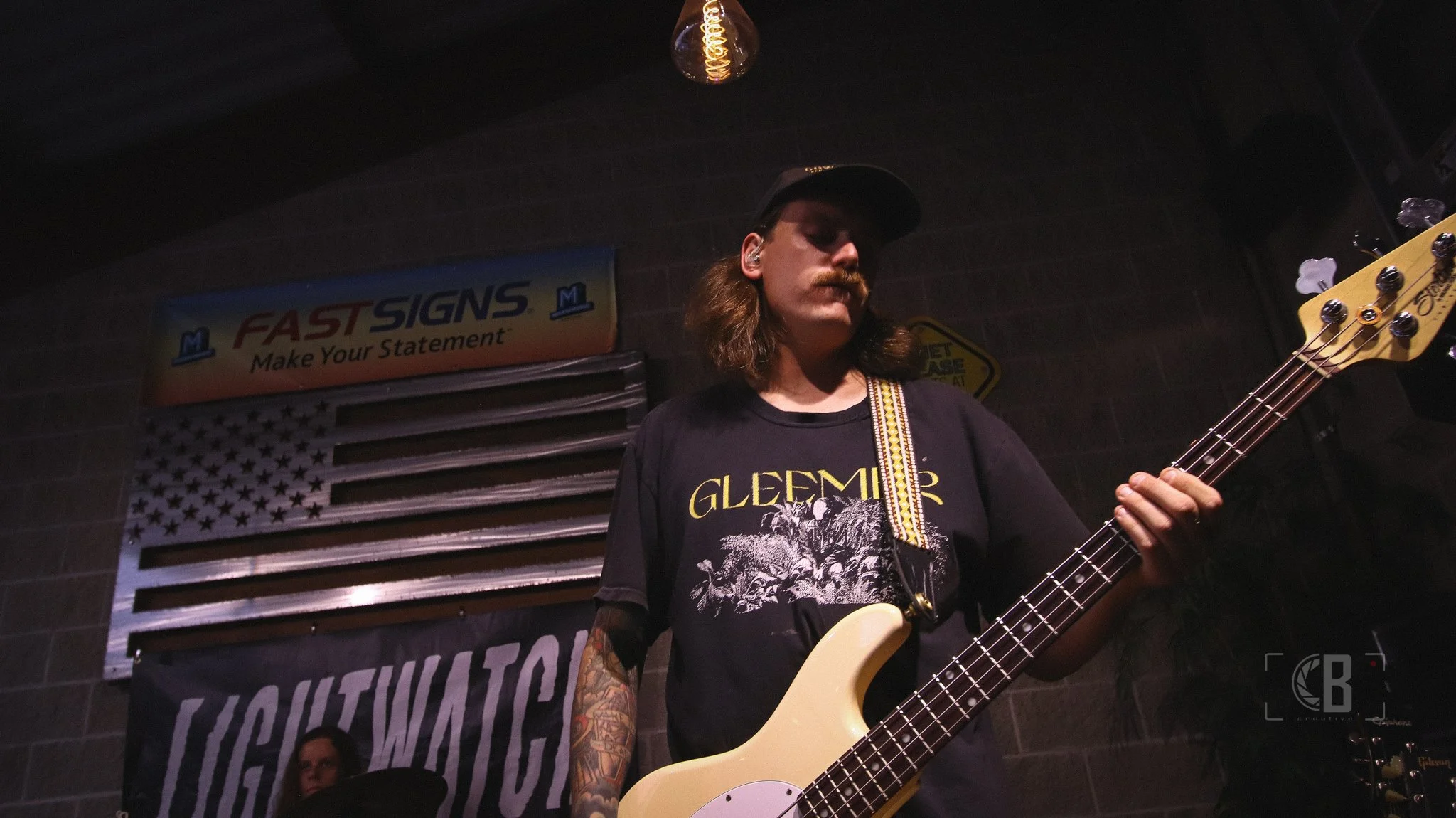 A man with long hair, mustache, tattoos, wearing a black T-shirt with a graphic and a black cap, playing a cream-colored electric bass guitar on stage. Behind him is an American flag and a sign that says 'FASTSIGNS Make Your Statement'.