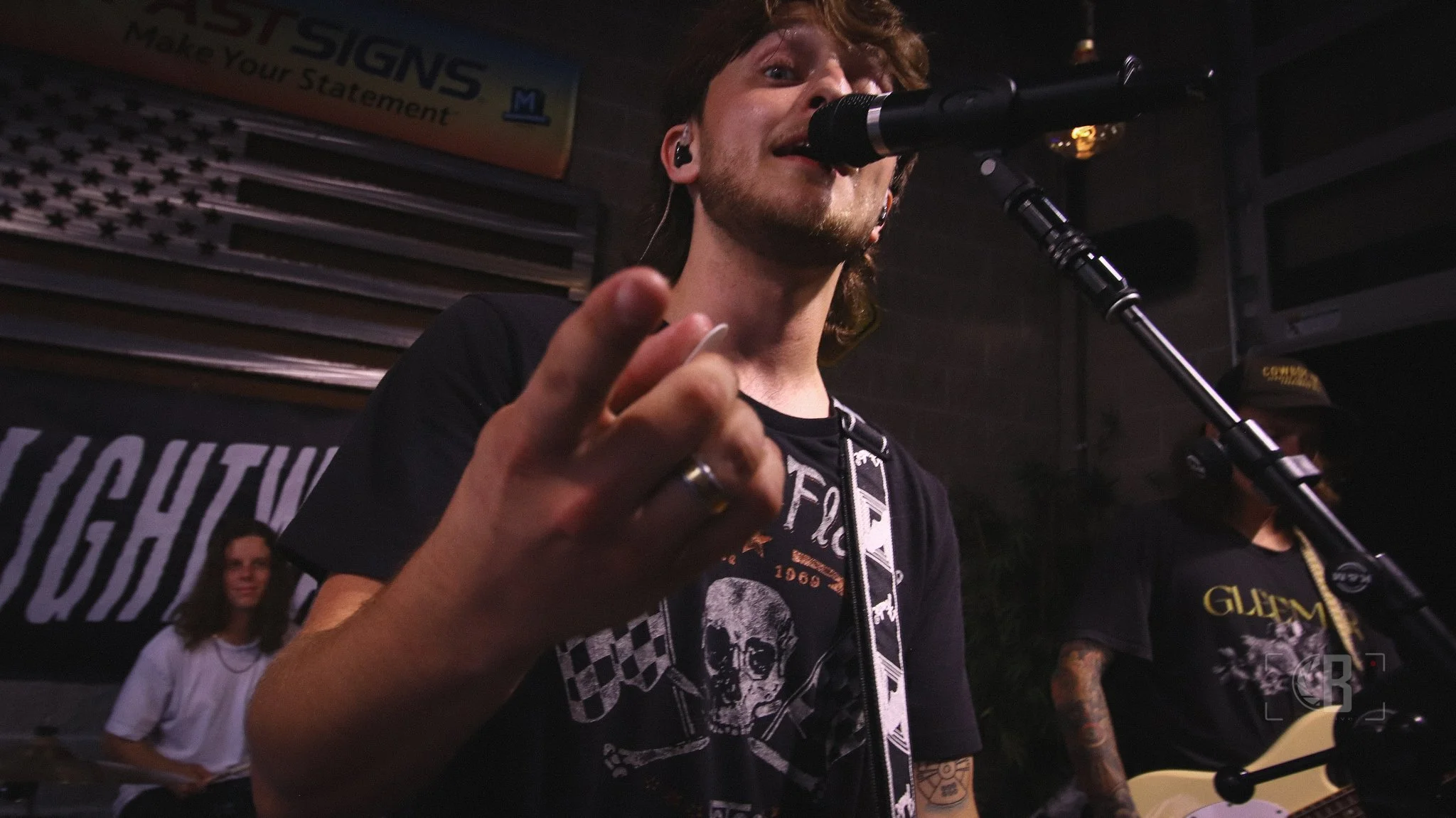 A male musician with curly hair and a beard singing into a microphone during a live performance, wearing a black t-shirt with a skull design, with a woman and another musician playing guitar in the background.