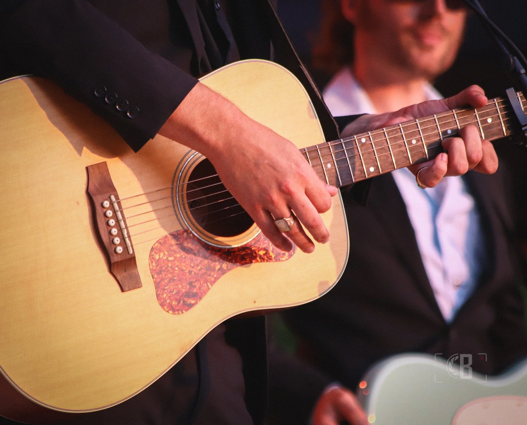 Person playing an acoustic guitar during a performance, with a second person visible in the background.
