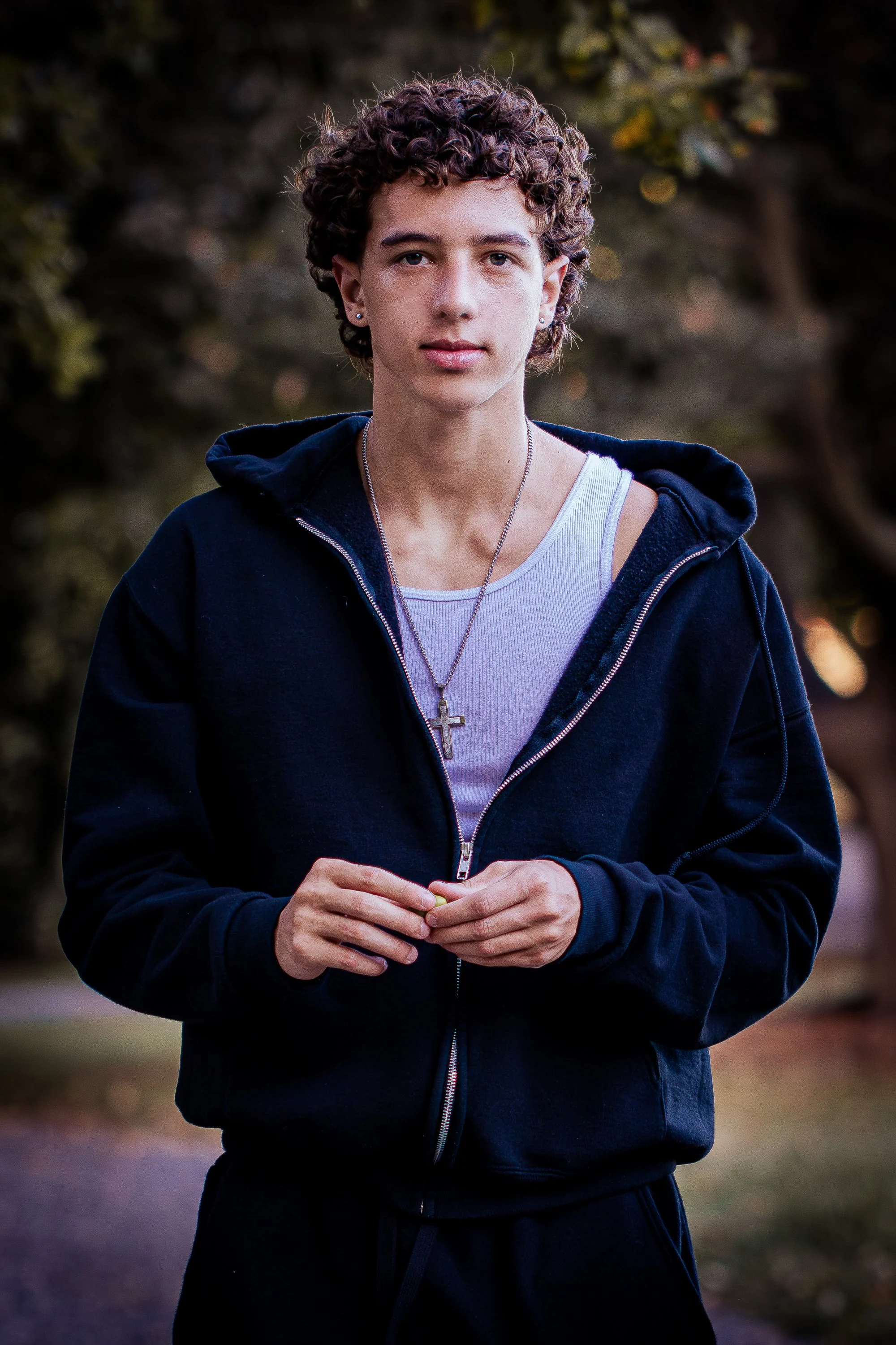 A young woman with curly brown hair and earrings, wearing a white tank top, a cross necklace, and a black zip-up hoodie, standing outdoors during dusk with a blurred background of trees and foliage.