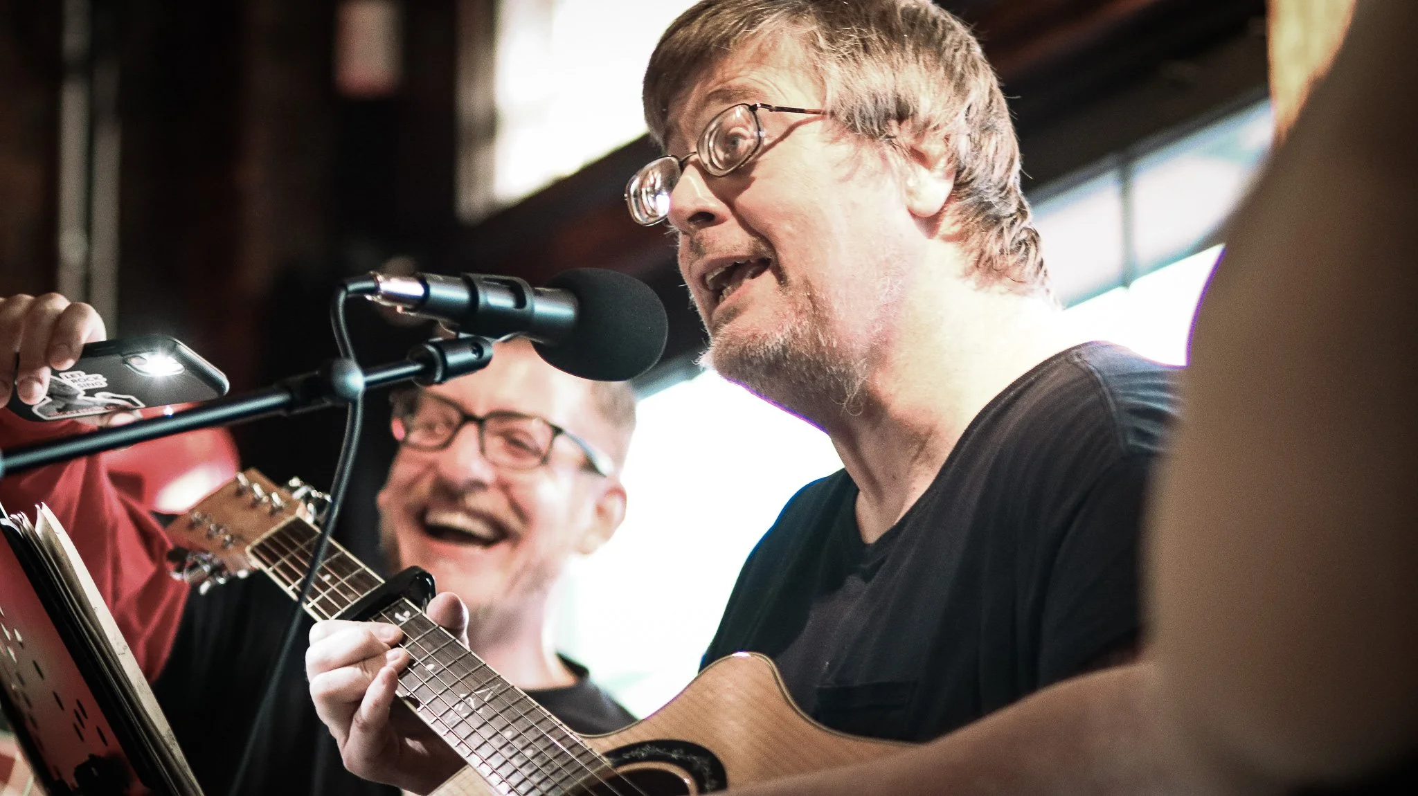 Two men singing and playing guitars at a live music performance, one man is wearing glasses and the other has a beard and glasses, with a microphone in front of them.