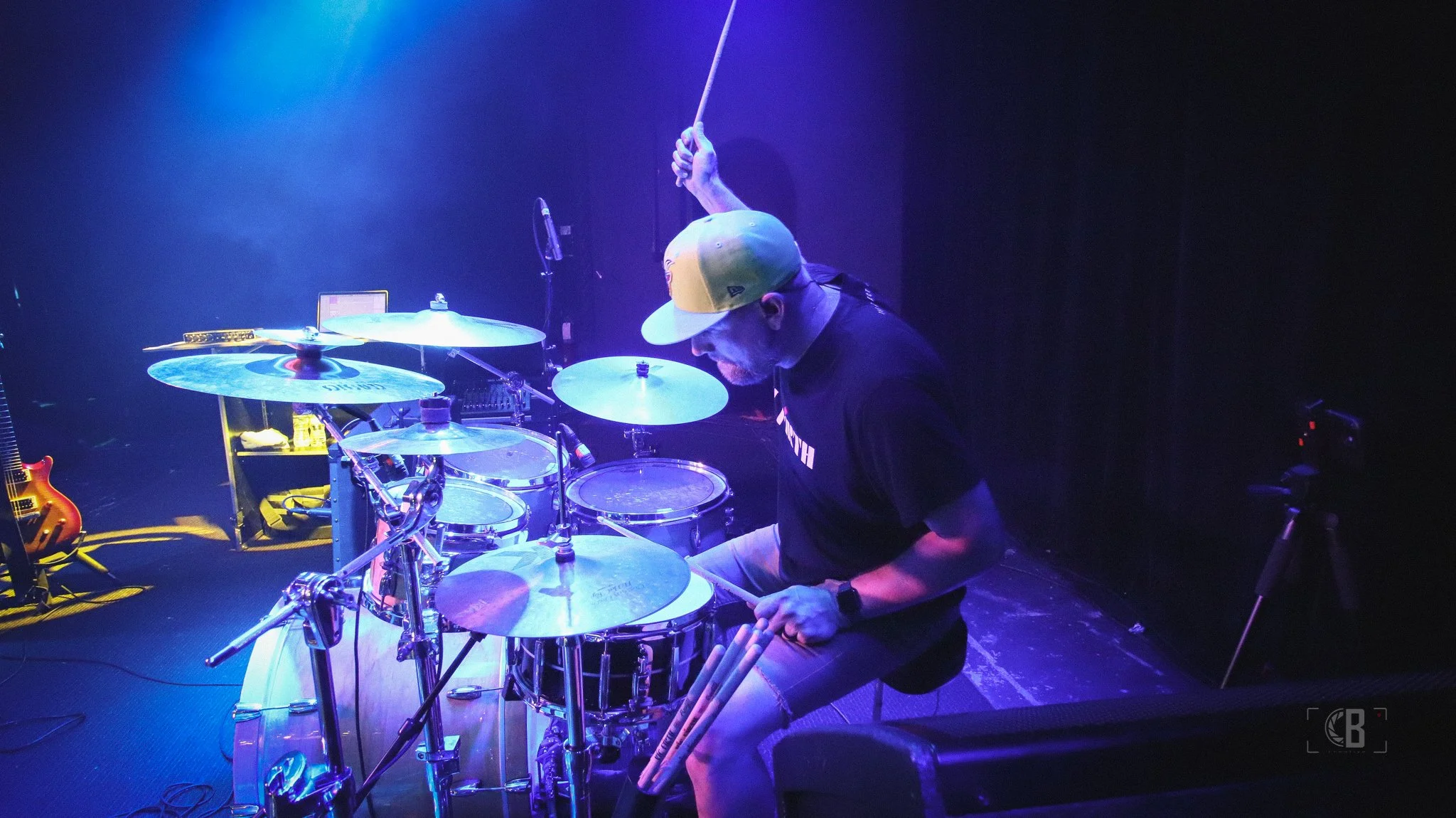 A man wearing a yellow and gray baseball cap, a black T-shirt, and shorts, playing a drum set on a dimly lit stage with purple and blue lighting.