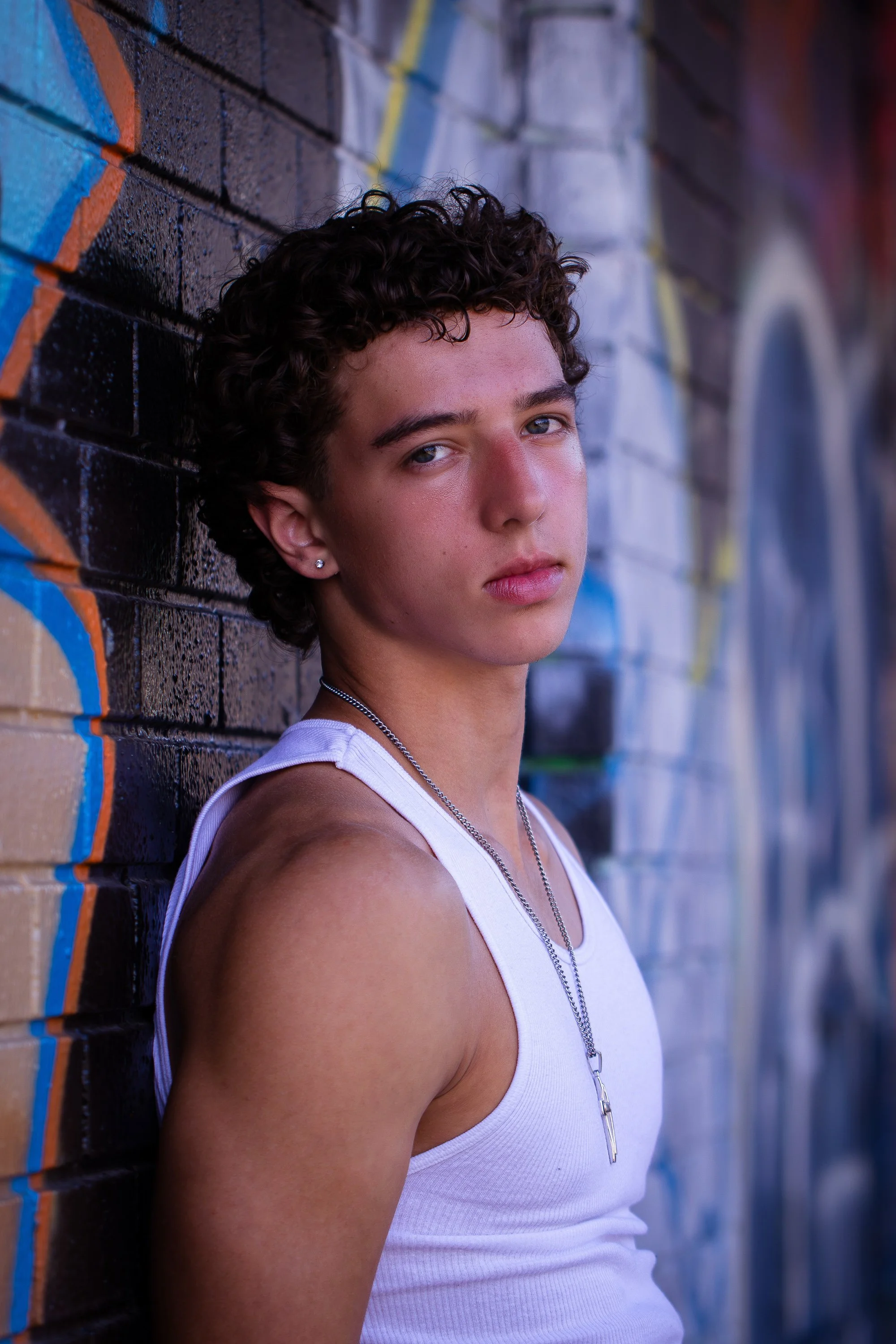 A young man with curly hair and earrings leaning against a colorful graffiti-covered brick wall, looking at the camera.