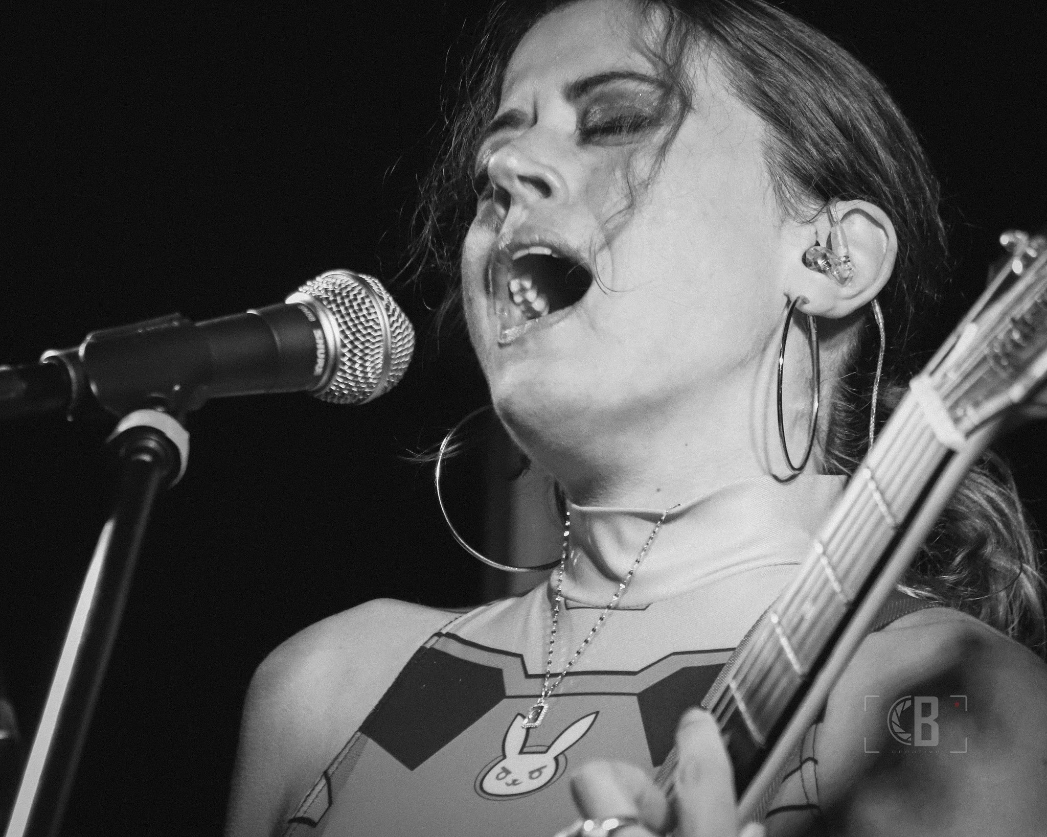 Black and white photo of a woman singing into a microphone while playing a guitar, with her eyes closed and mouth open, wearing hoop earrings, a necklace, and a tank top with a Bunny design.