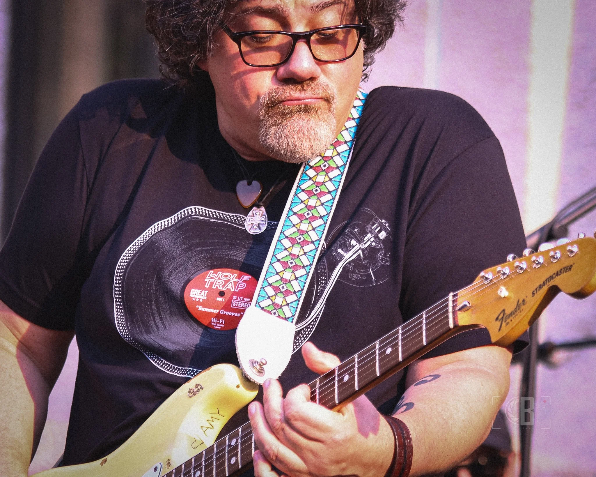 A man with curly gray hair, glasses, and a beard, playing an electric guitar. He is wearing a black T-shirt with a vinyl record print and a colorful guitar strap.