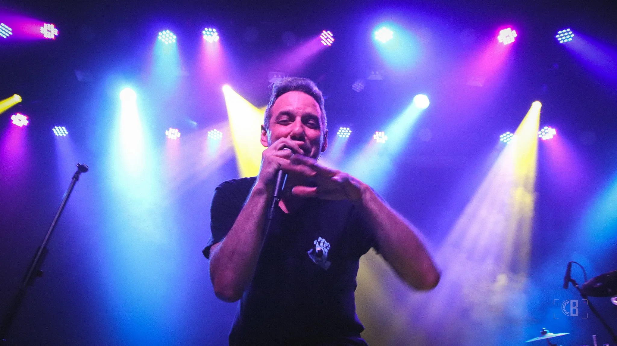 A man performing on stage with colorful lights shining behind him, holding a microphone and making a gesture with his right hand.