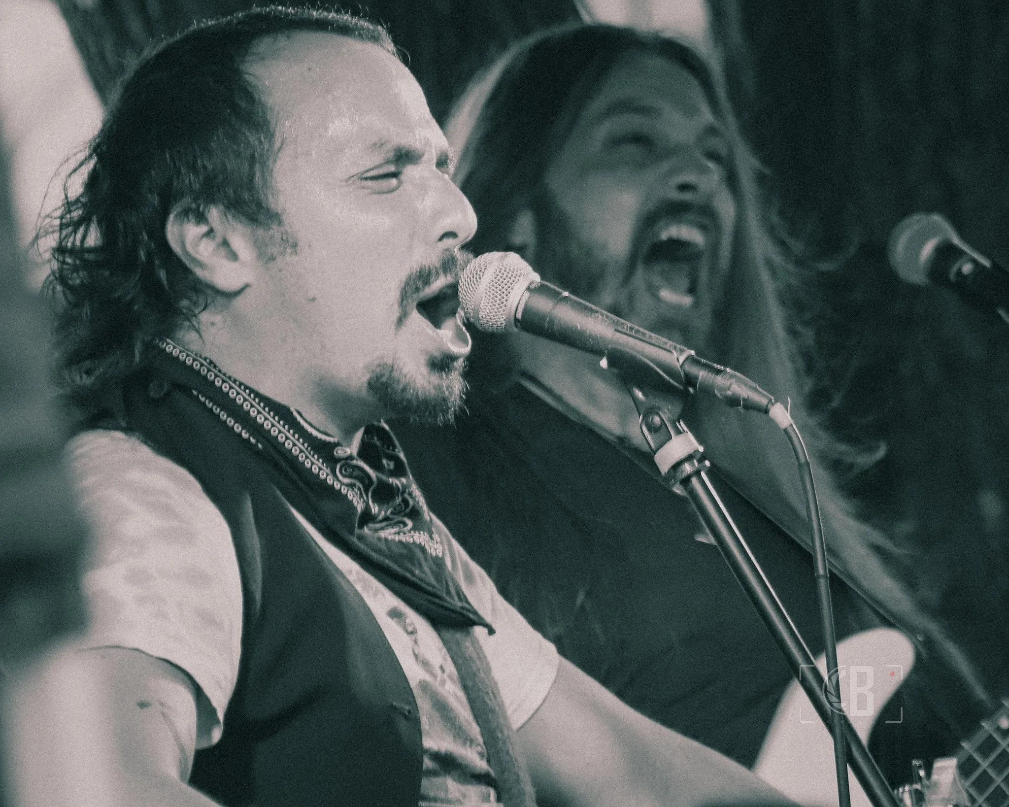 Two male musicians singing into microphones, one playing guitar, performance in black and white.