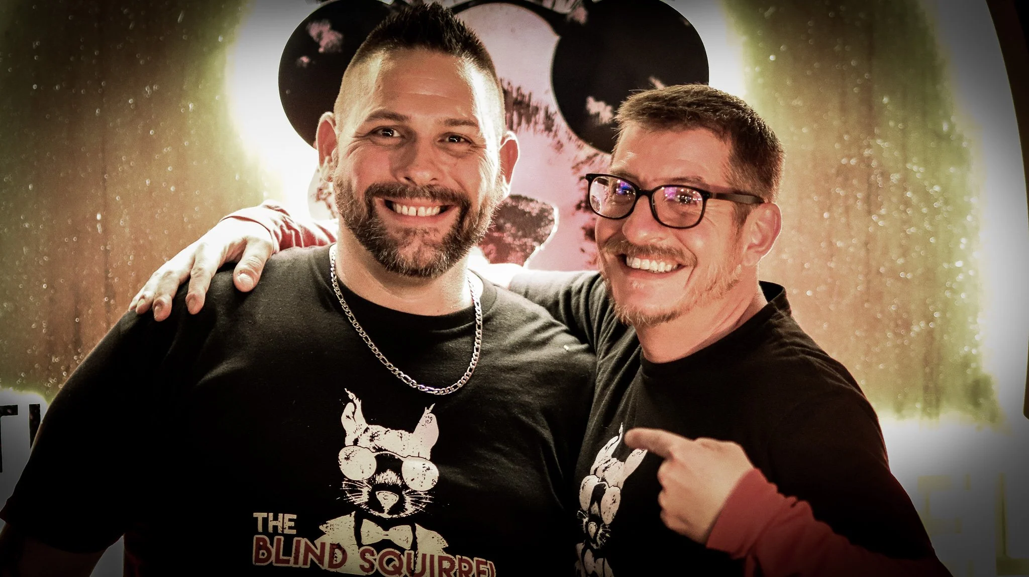 Two smiling men with beards and glasses wearing black T-shirts with a graphic of a cat with sunglasses and the text 'The Blind Squirrel', standing close together with their arms around each other in front of a black and white skull artwork.
