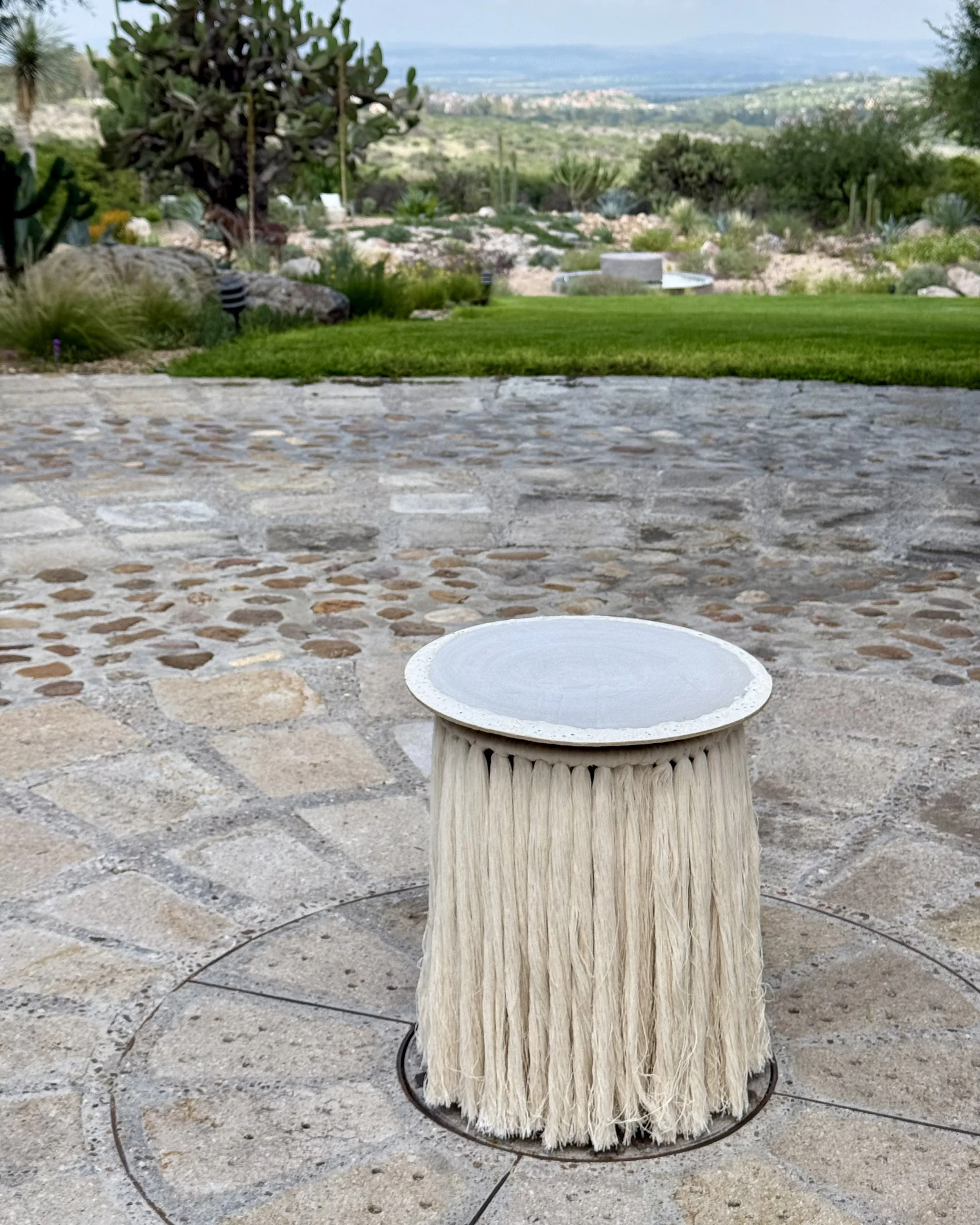 A decorative outdoor table with a cylindrical base made of textured light-colored material, and a round, flat top with a slightly raised edge, set on a stone patio.