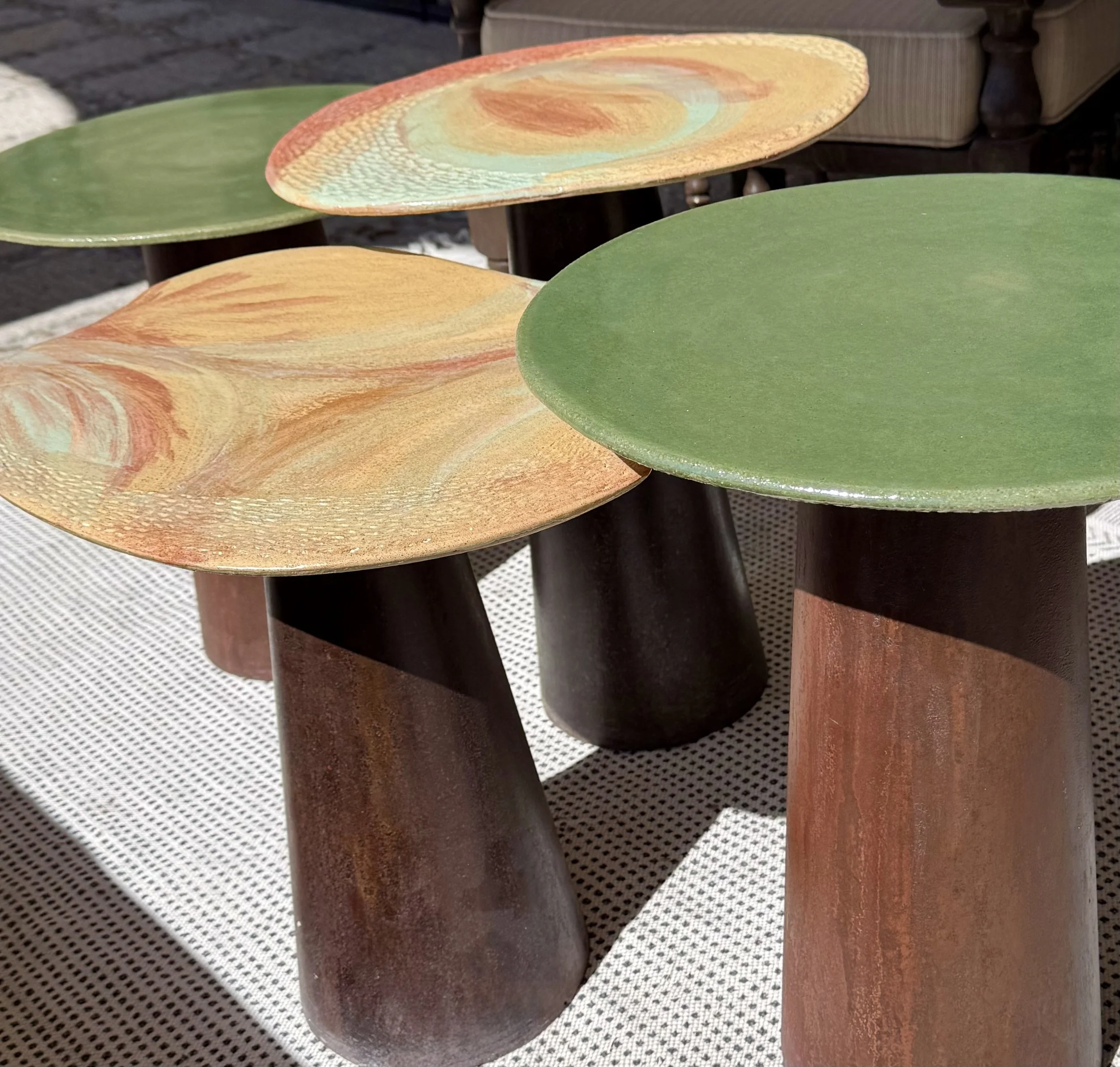 Close-up of colorful wooden stools with round, flat tops and cylindrical bases, in shades of green, orange, and brown, placed on a patterned surface.