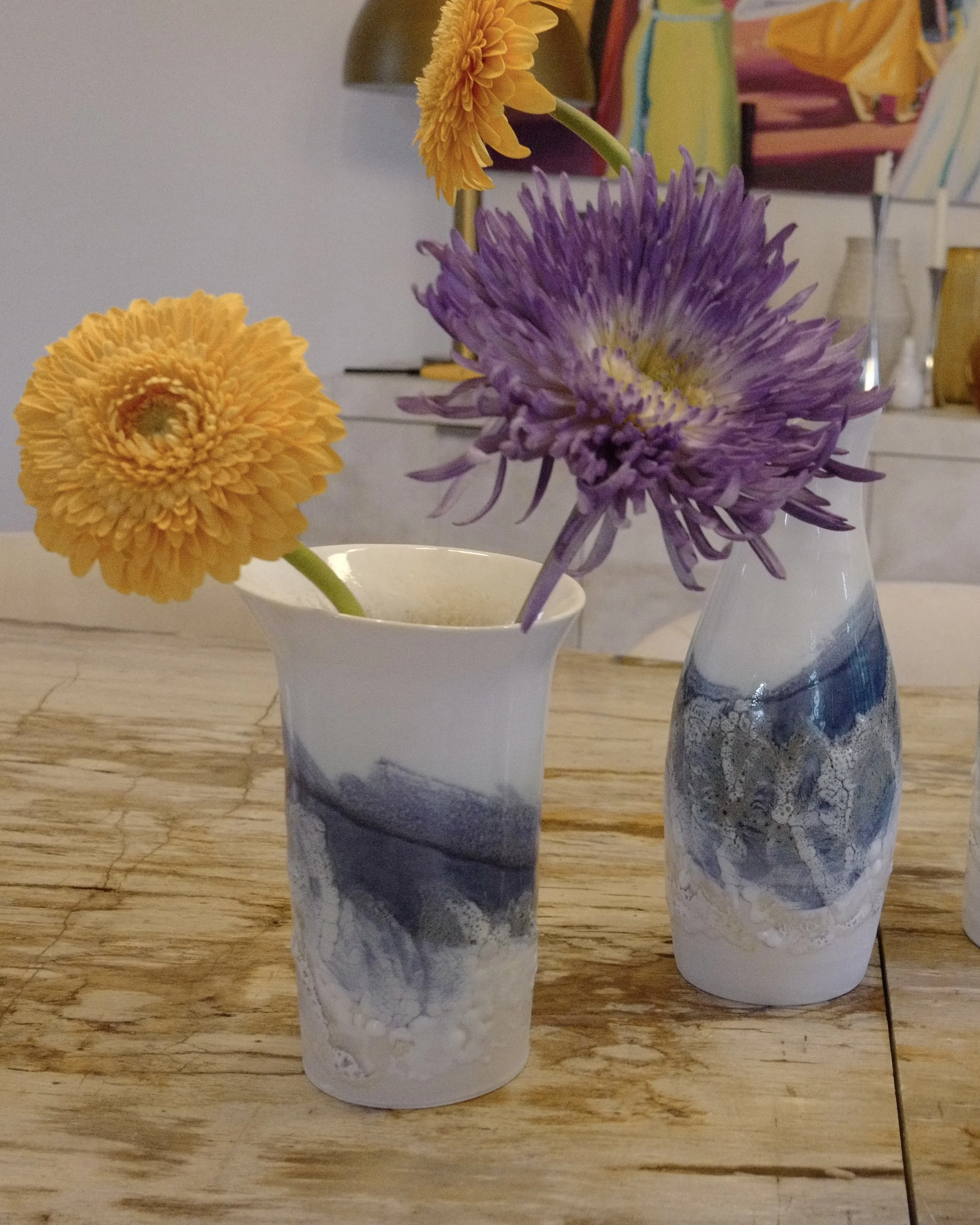Close-up of a purple and yellow flower in a white and blue marbled ceramic vase on a wooden table, with another similar vase in the background containing yellow flowers.