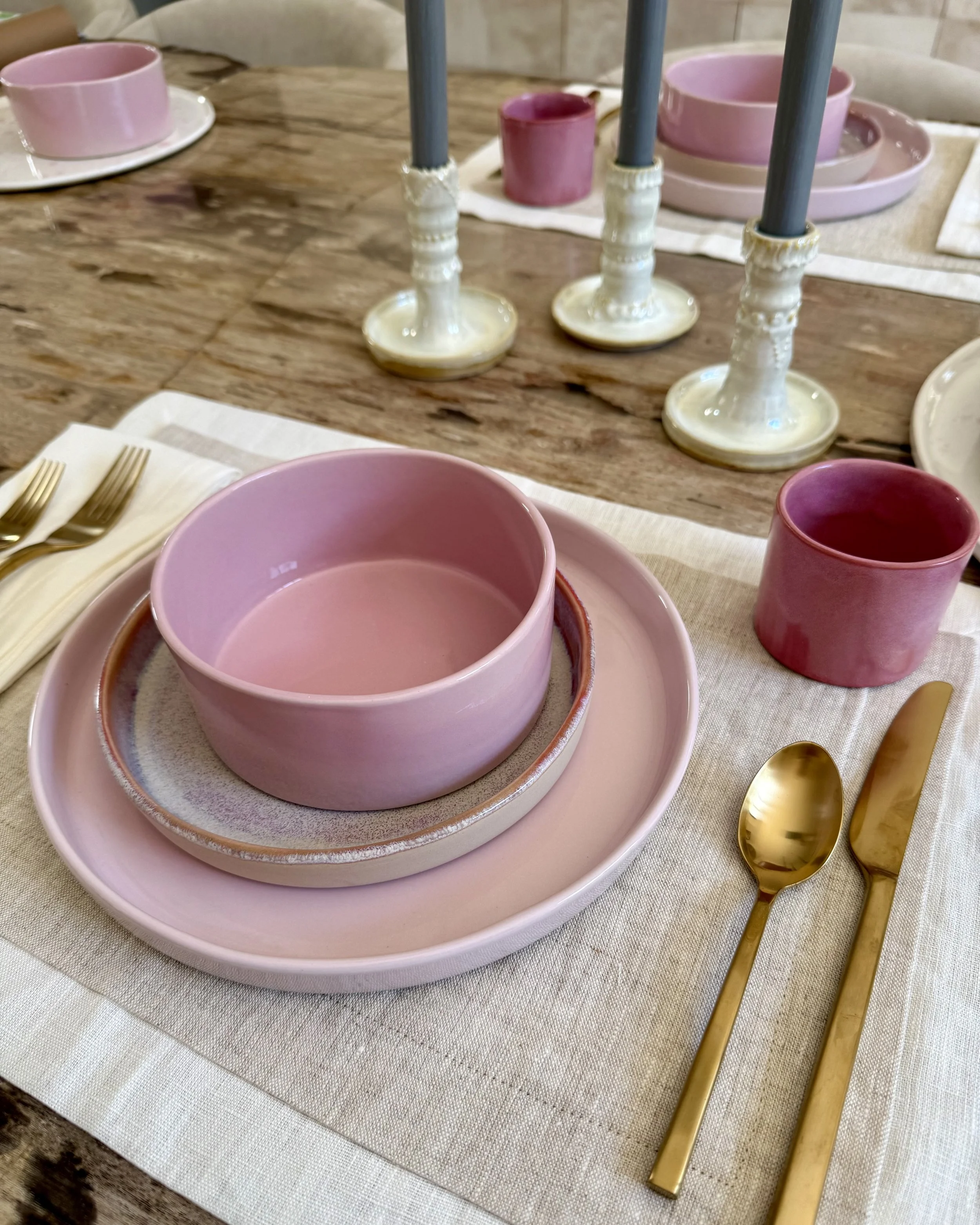 A table setting with pink and gold dishware, including pink bowls, pink cups, and gold cutlery, on a cream-colored tablecloth with a natural wooden table surface visible.