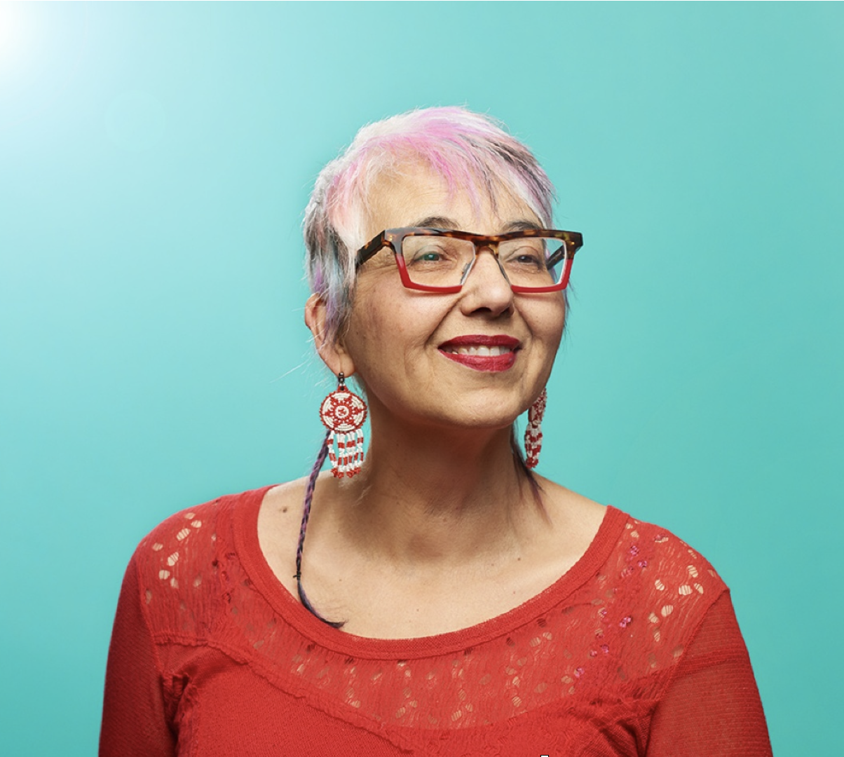 An older woman with short, multi-colored hair, wearing red and black glasses, red lipstick, and earrings, smiling against a solid teal background.