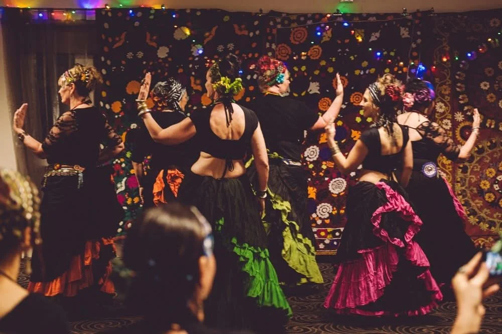 Six women of Temple Rain Dance performing a folkloric dance in colorful skirts and blouses, with a floral tapestry background and string lights overhead.