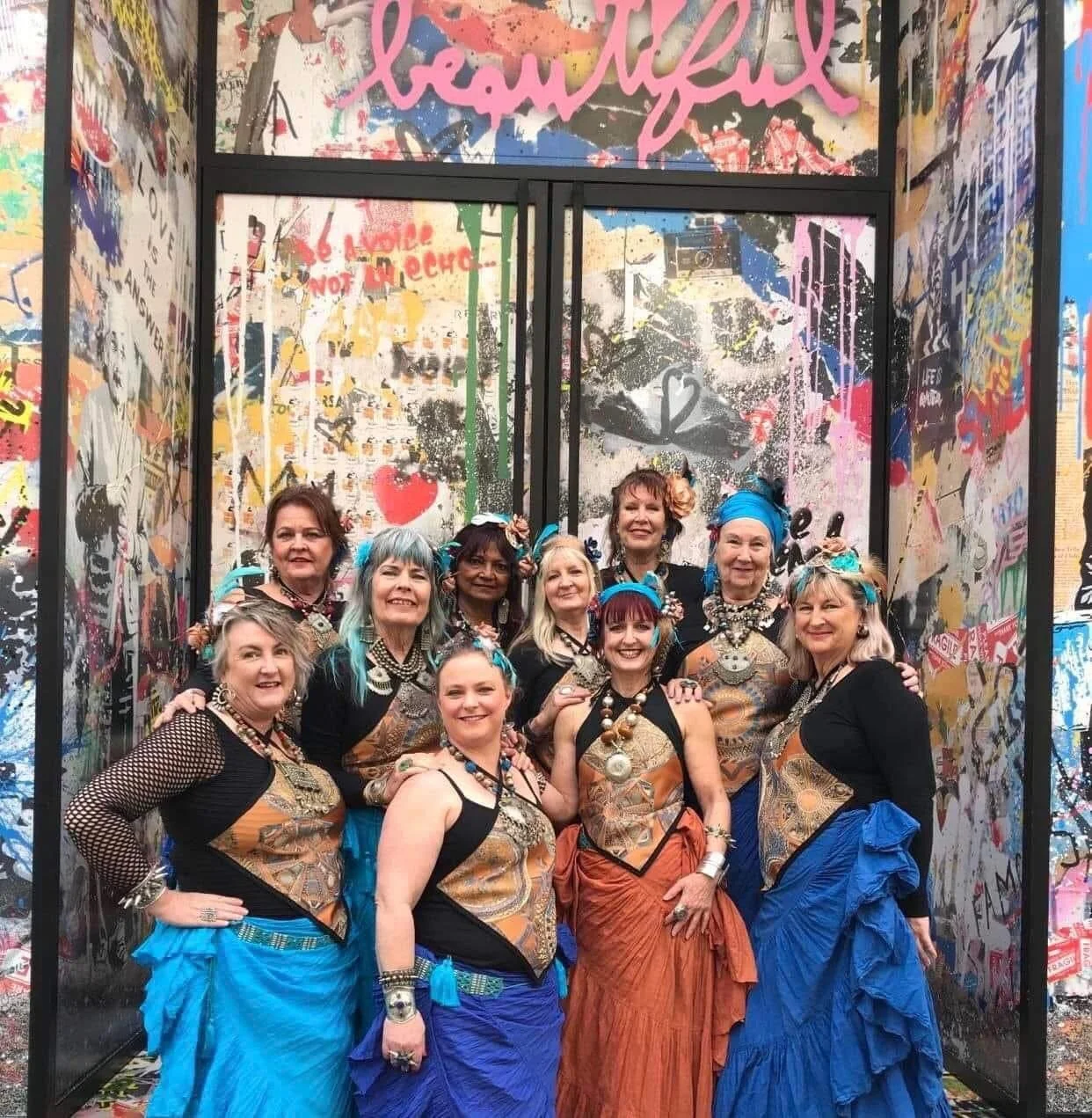 A group of nine women dressed in bohemian style outfits with skirts, jewelry, and colorful head wraps, posing together inside an art installation with graffiti and colorful artwork on the walls and the word 'beautiful' written in pink on the top.