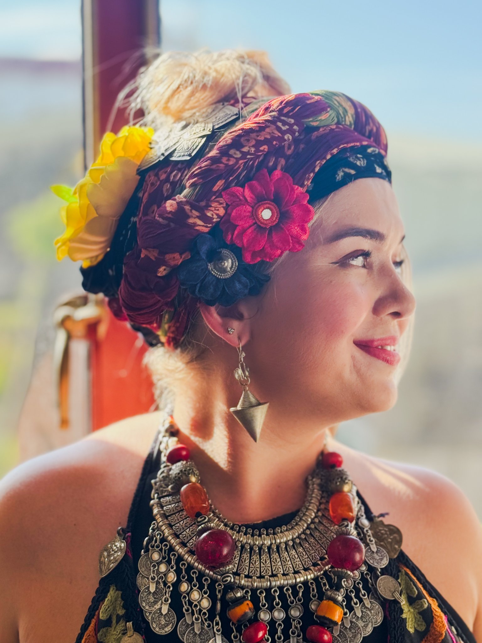 A Global Caravan dancer - Jade - wearing a colorful headscarf with flowers and traditional jewelry, standing near a window with a blurred outdoor background.