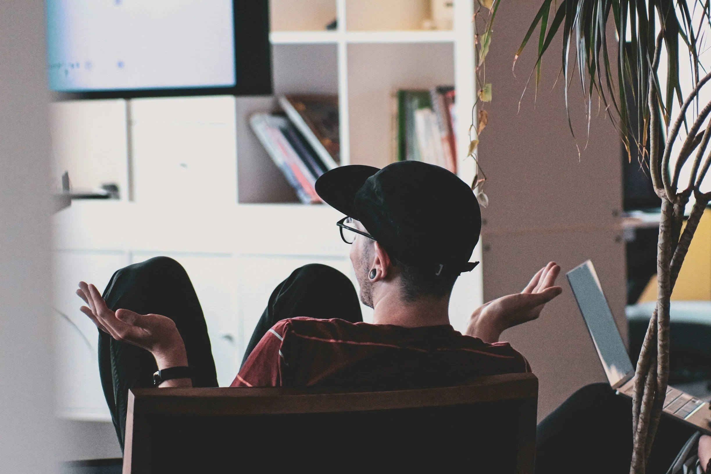 Person sitting in a chair, wearing a black cap and glasses, gesturing with hands, surrounded by bookshelves and a potted plant. Representing an individual therapy session with an Affirmative Minds Counselling Therapy professional.