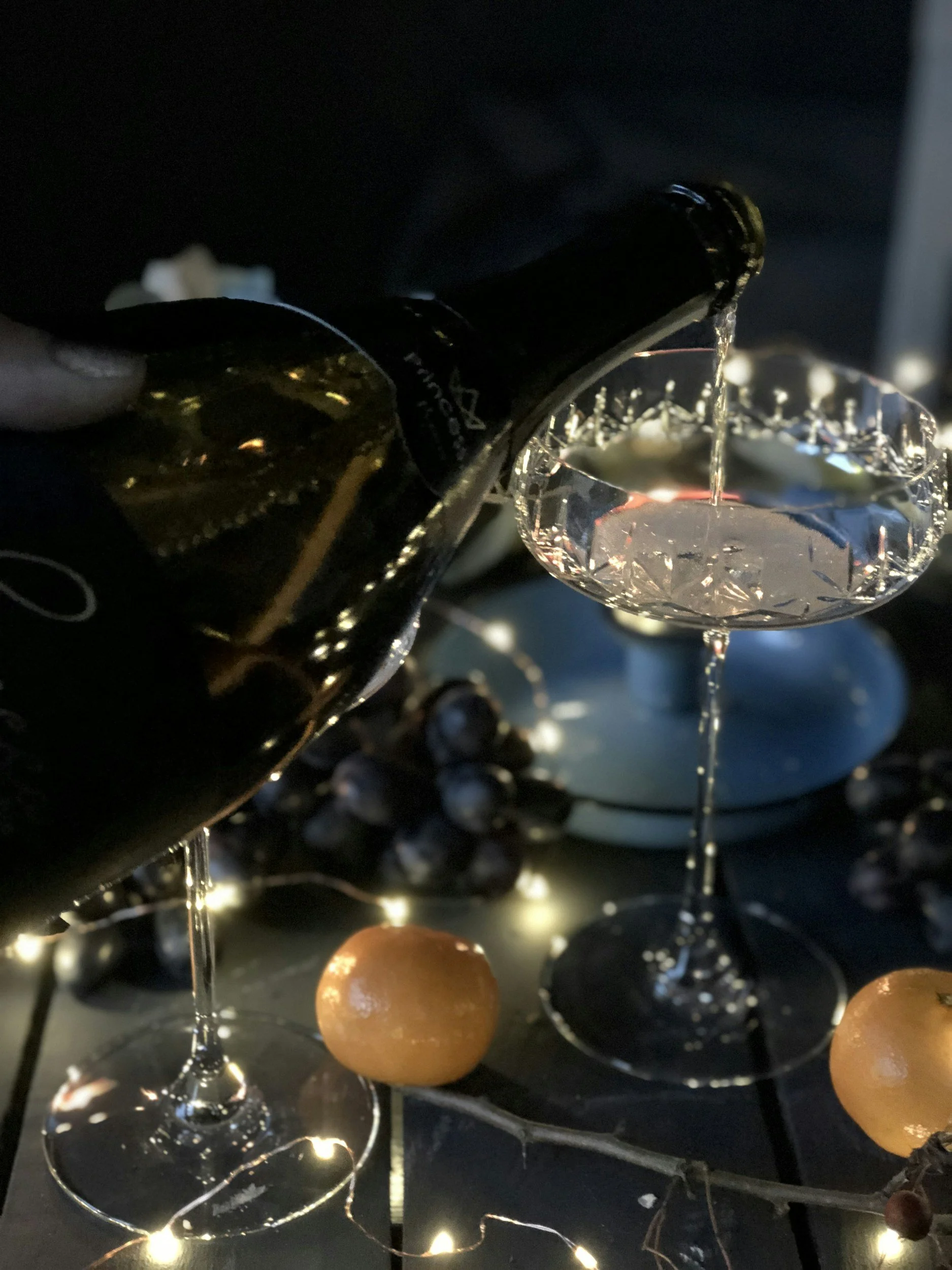 Pouring champagne into a coupe glass, with a dark background decorated with grapes, mandarins, and fairy lights.