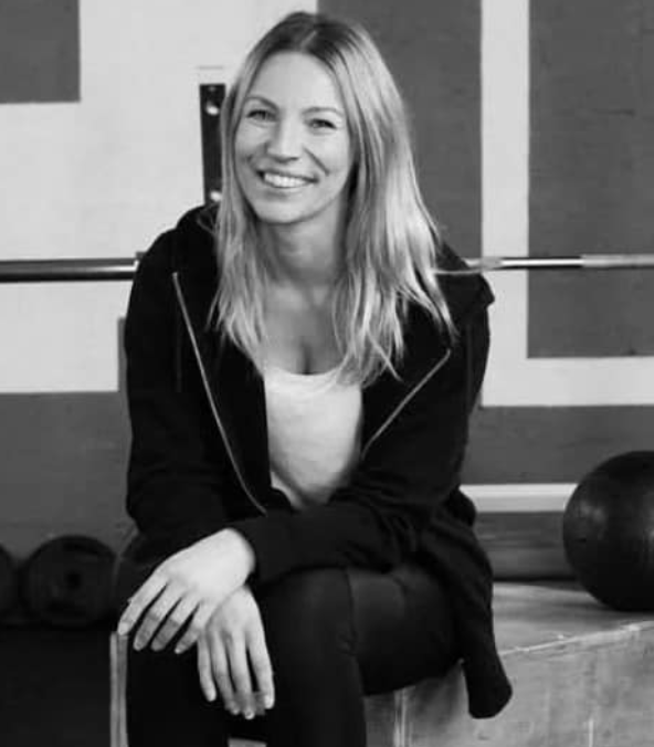 A smiling woman with long blonde hair sitting on a wooden box in a gym, wearing a black hoodie and white top, surrounded by gym equipment.