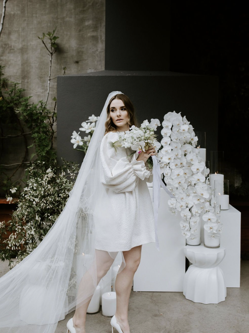 A bride in a white dress and veil holding a bouquet of white flowers, standing in front of a modern floral display with white orchids and candles at The French Cafe by Together Journal
