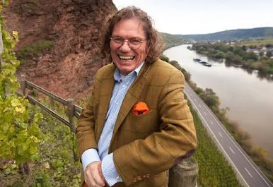 A smiling man with curly hair and glasses standing outdoors near a river, with a road and green trees in the background.