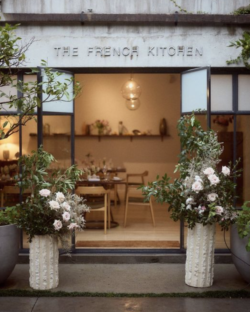 Entrance to a restaurant called 'The French Kitchen' with open doors, floral arrangements in white vases on either side, and a cozy, warmly lit interior with wooden tables and chairs.