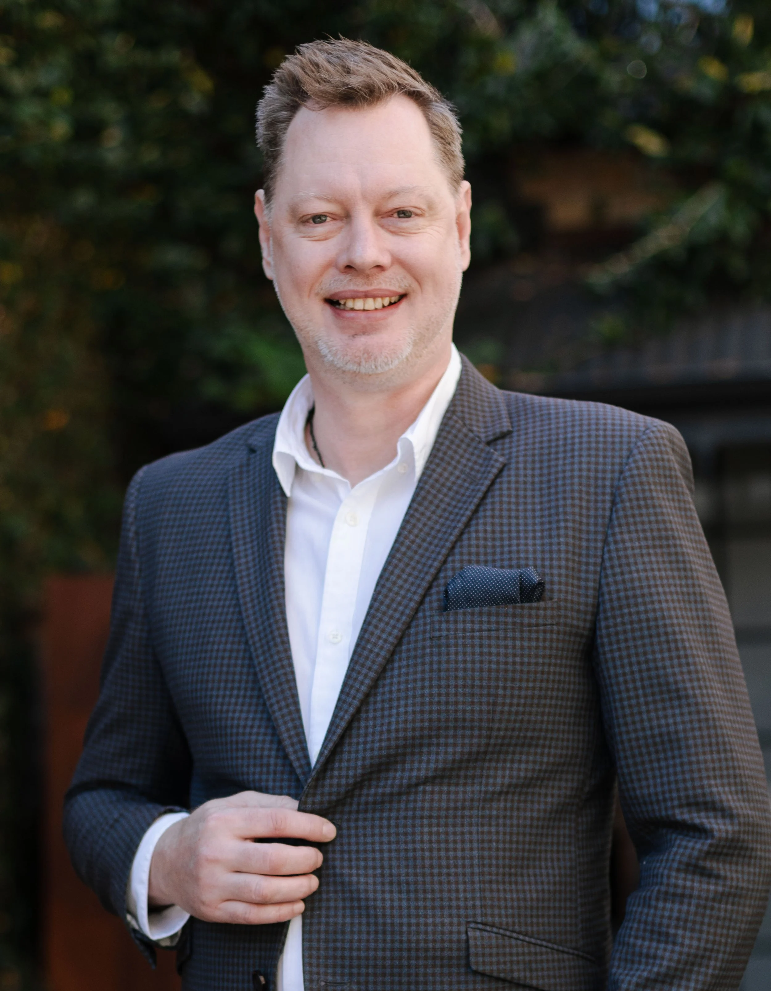 Matthew Aitchison, wearing a navy blazer over a checkered shirt, smiling and standing in front of green leafy plants.