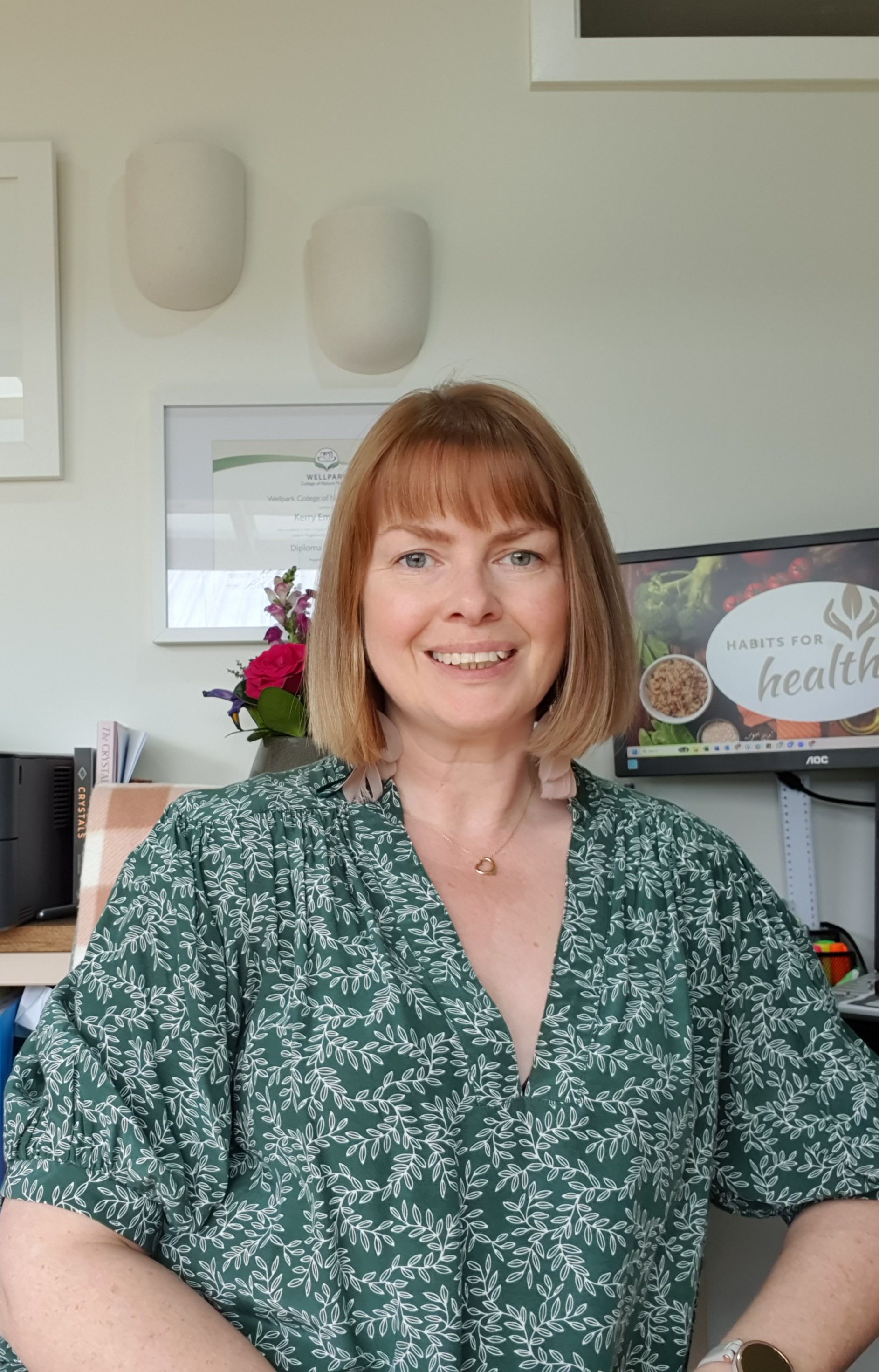 A woman with shoulder-length red hair wearing a green-patterned blouse, sitting at a desk in an office. Behind her, a computer monitor displays a presentation titled 'Habits for Health.' There are framed certificates, a flower arrangement, and office supplies visible in the background.
