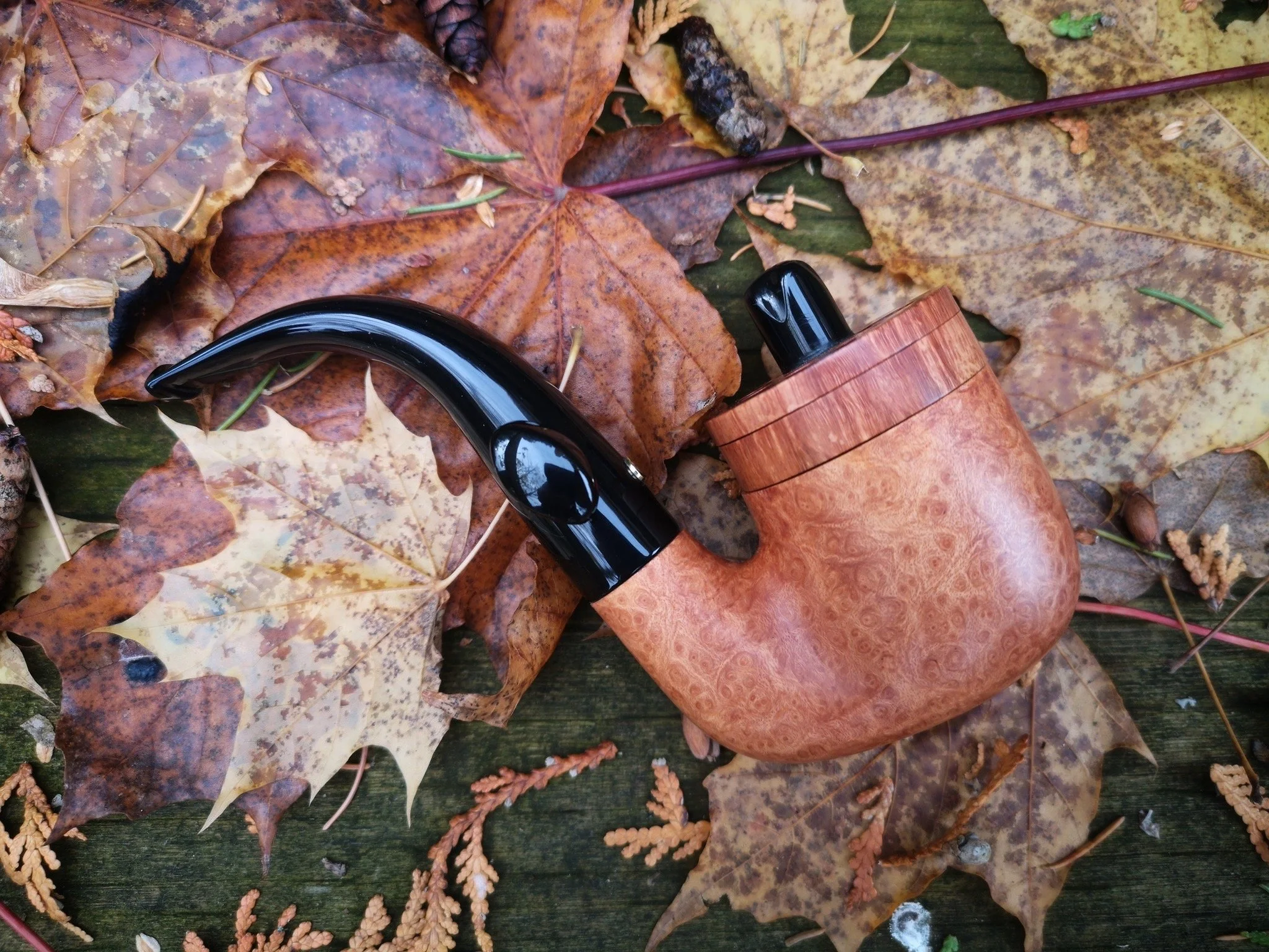 A wooden smoking pipe with a black mouthpiece lying on fallen autumn leaves on a wooden surface.