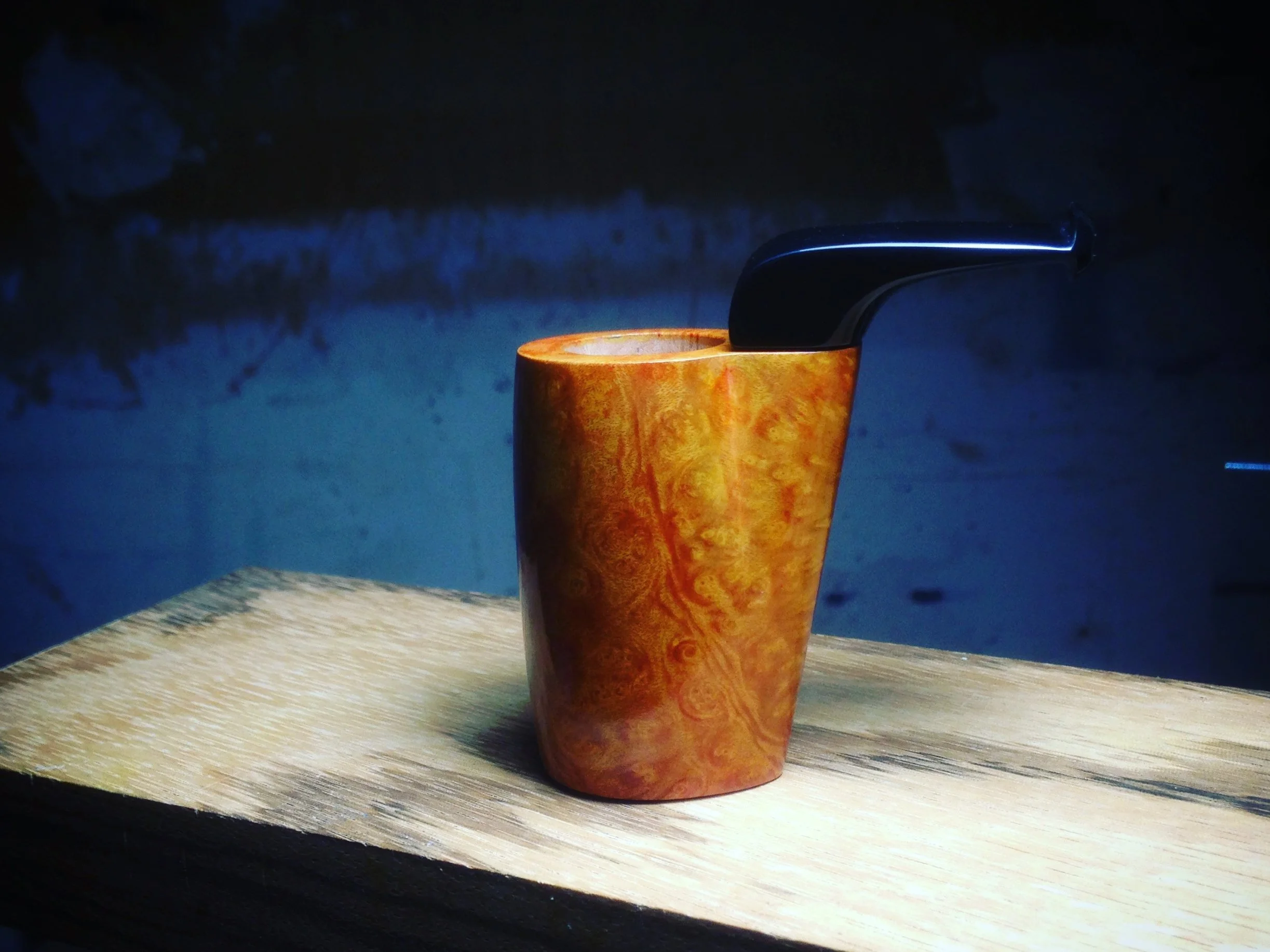 A smoking pocket pipe with a black stem and a orange-brown bowl with a swirl pattern, on a wooden surface with a dark background.