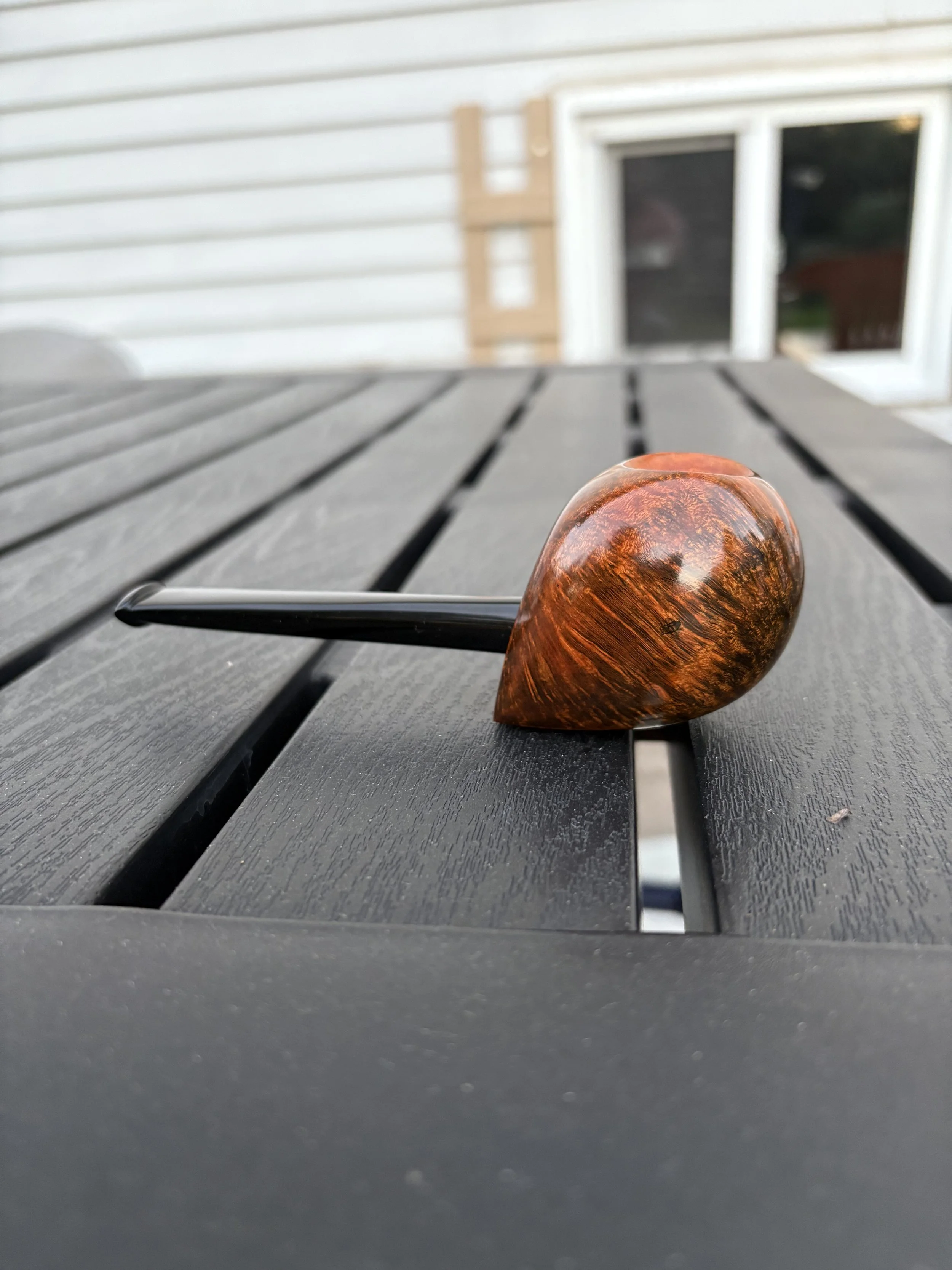 A freehand smoking pipe with a brown, glossy, wooden bowl and a black stem resting on a black wooden outdoor table.