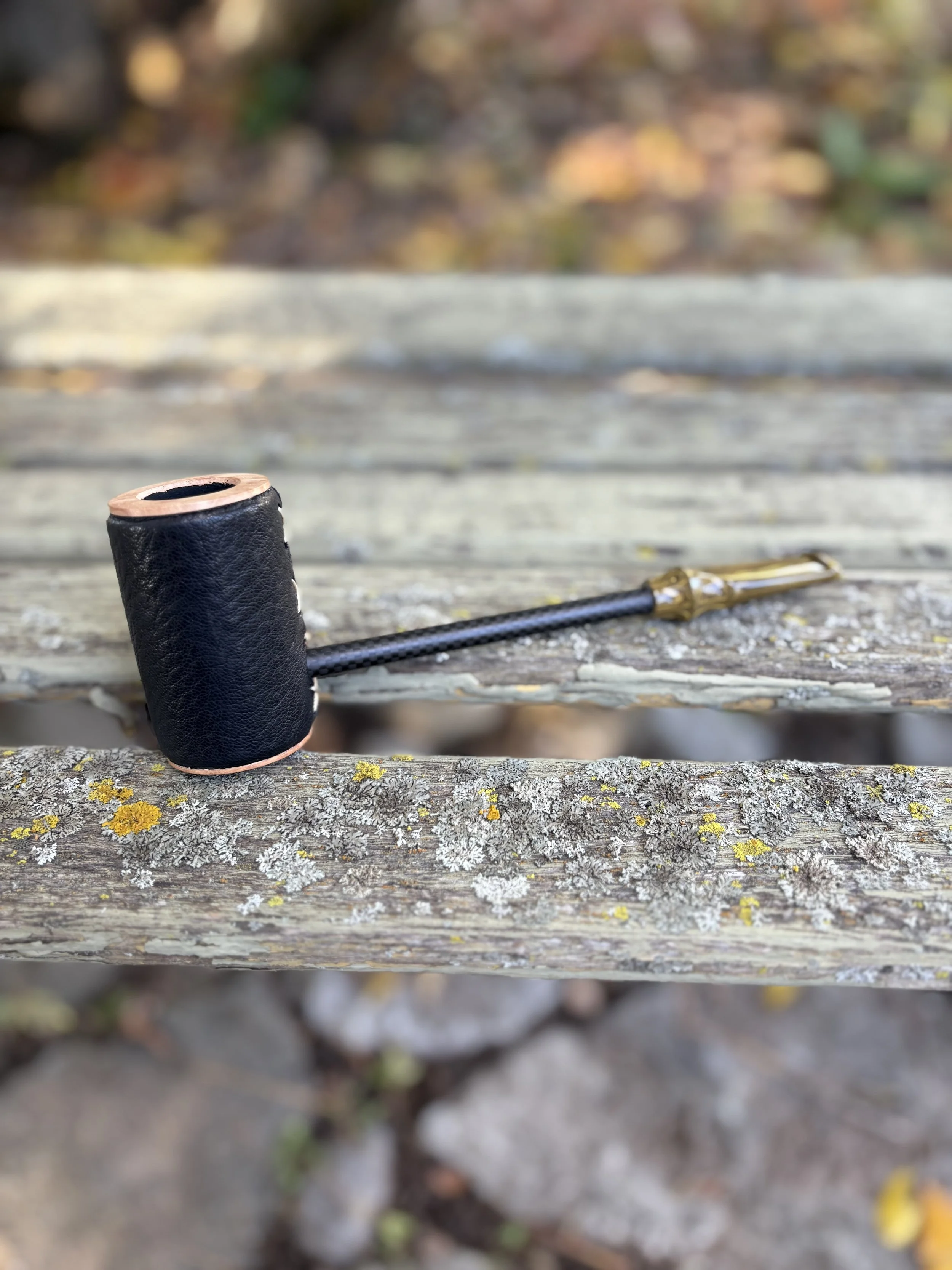 A close-up of a leather-wrapped poker smoking pipe with a black leather wrap and a long, gold-colored stem resting on a weathered wooden railing outdoors.