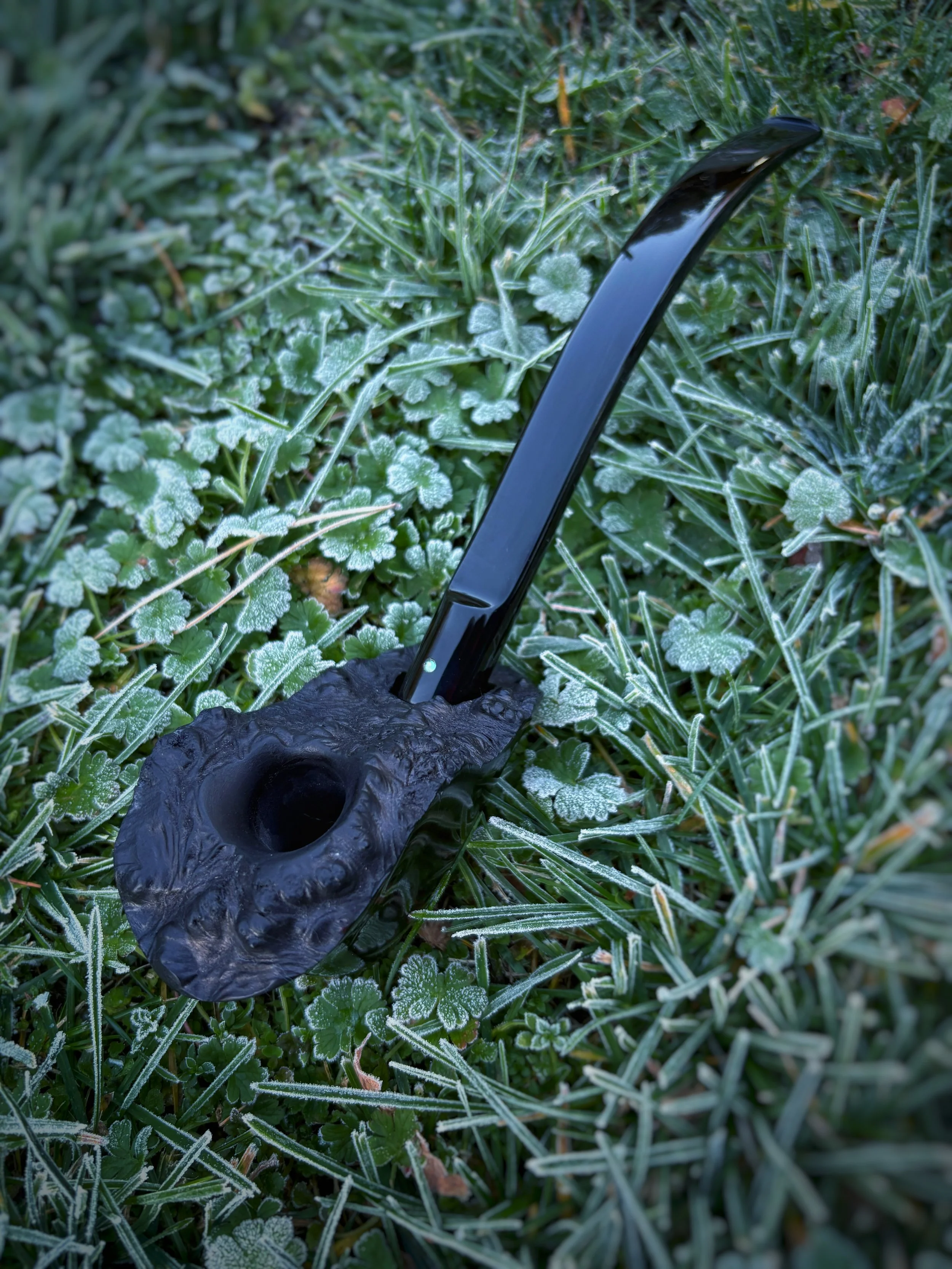 A Freehand smoking pipe with a black, textured bowl and a shiny black stem lying on frosty green grass with small leaves.