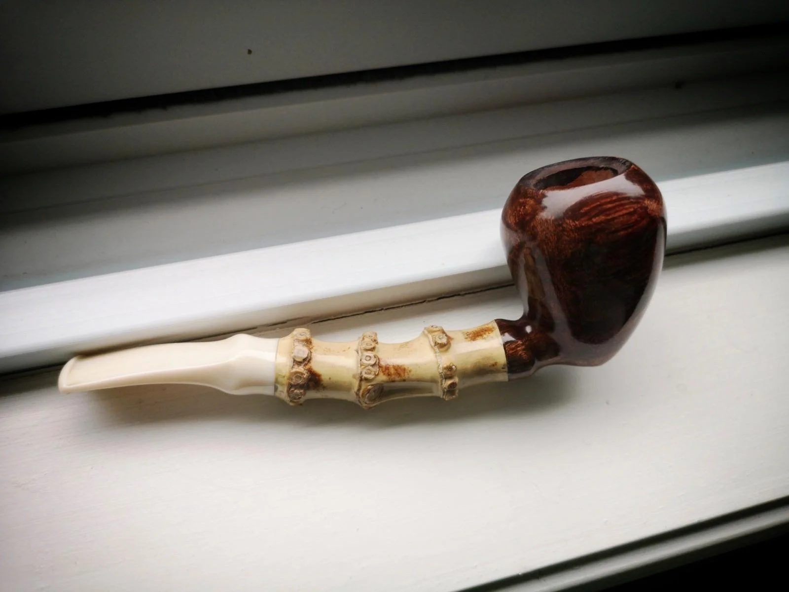 An acorn smoking pipe with a dark wooden bowl and a cream-colored stem lying on a white window sill