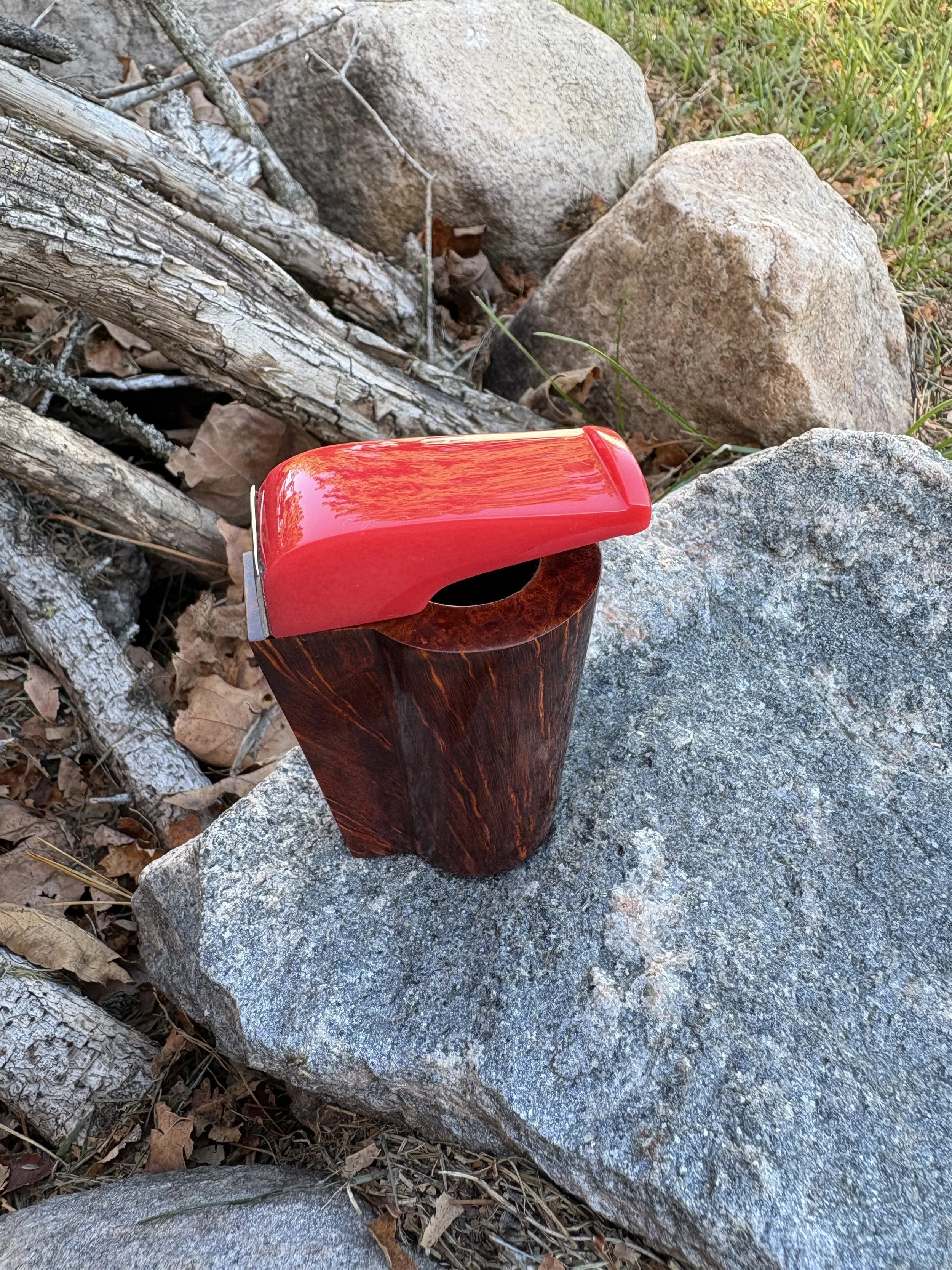 A briar pocket pipe lying on a large flat rock outdoors surrounded by rocks, dry leaves, and twigs.