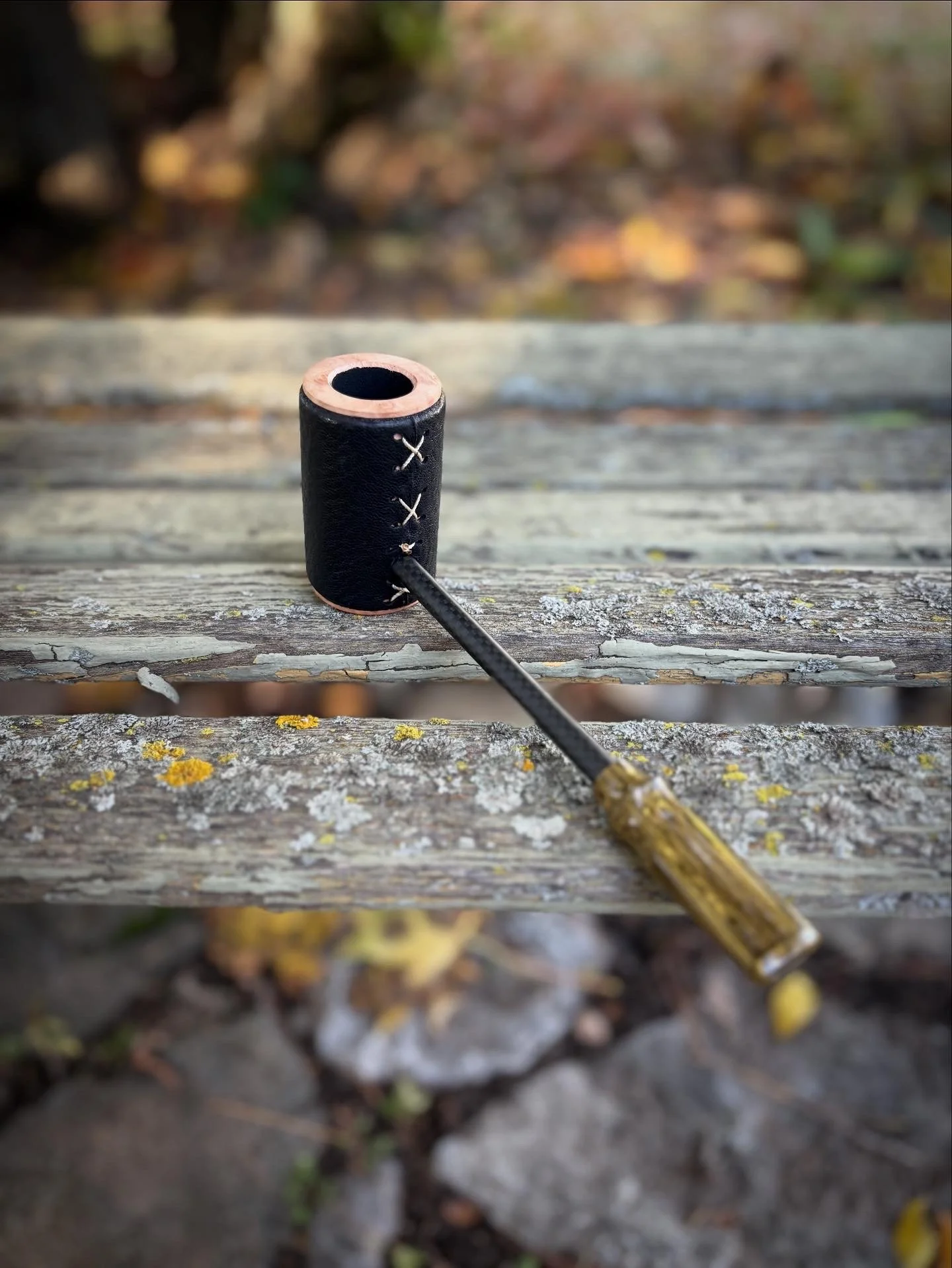 A leather-wrapped poker smoking pipe with a black leather-like cover and wooden handle resting on a weathered wooden bench outdoors with fallen leaves.