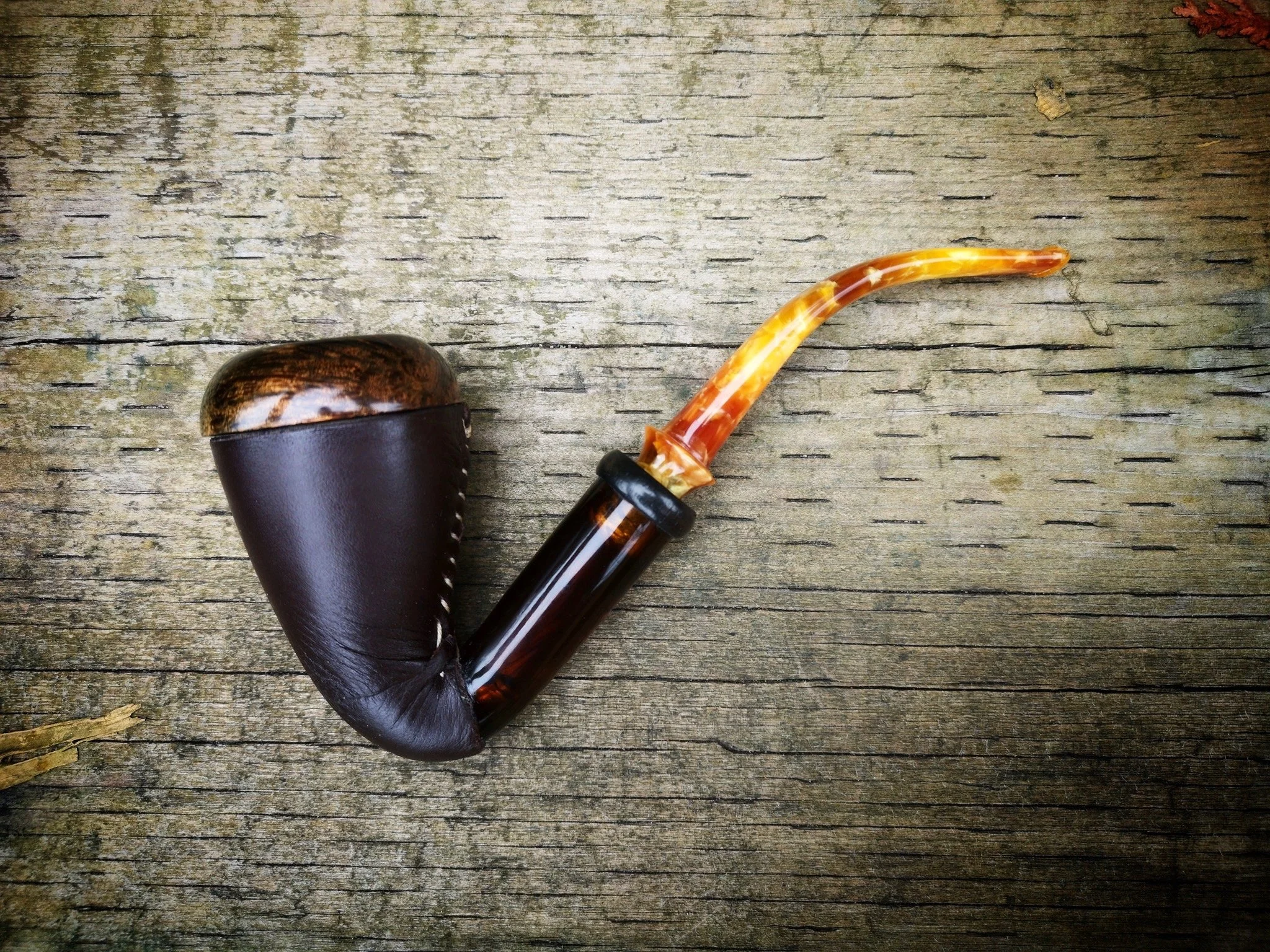 A  Calabash smoking pipe with a leather and wood bowl, a black mouthpiece, and a curved amber-colored stem resting on a weathered wooden surface.