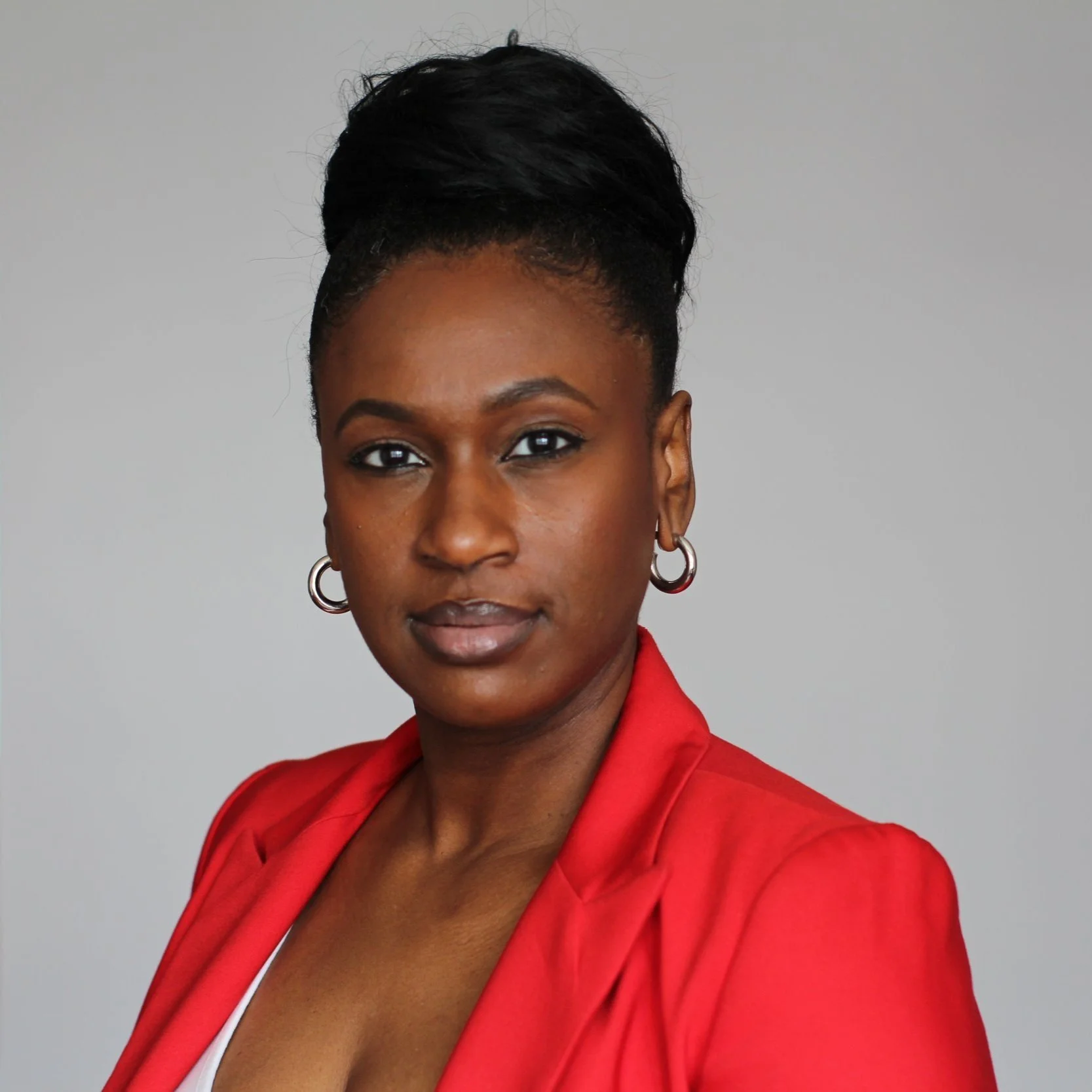 A professional portrait of a woman with dark skin, short black hair styled up, wearing a red blazer, hoop earrings, and neutral makeup, posing against a light gray background.