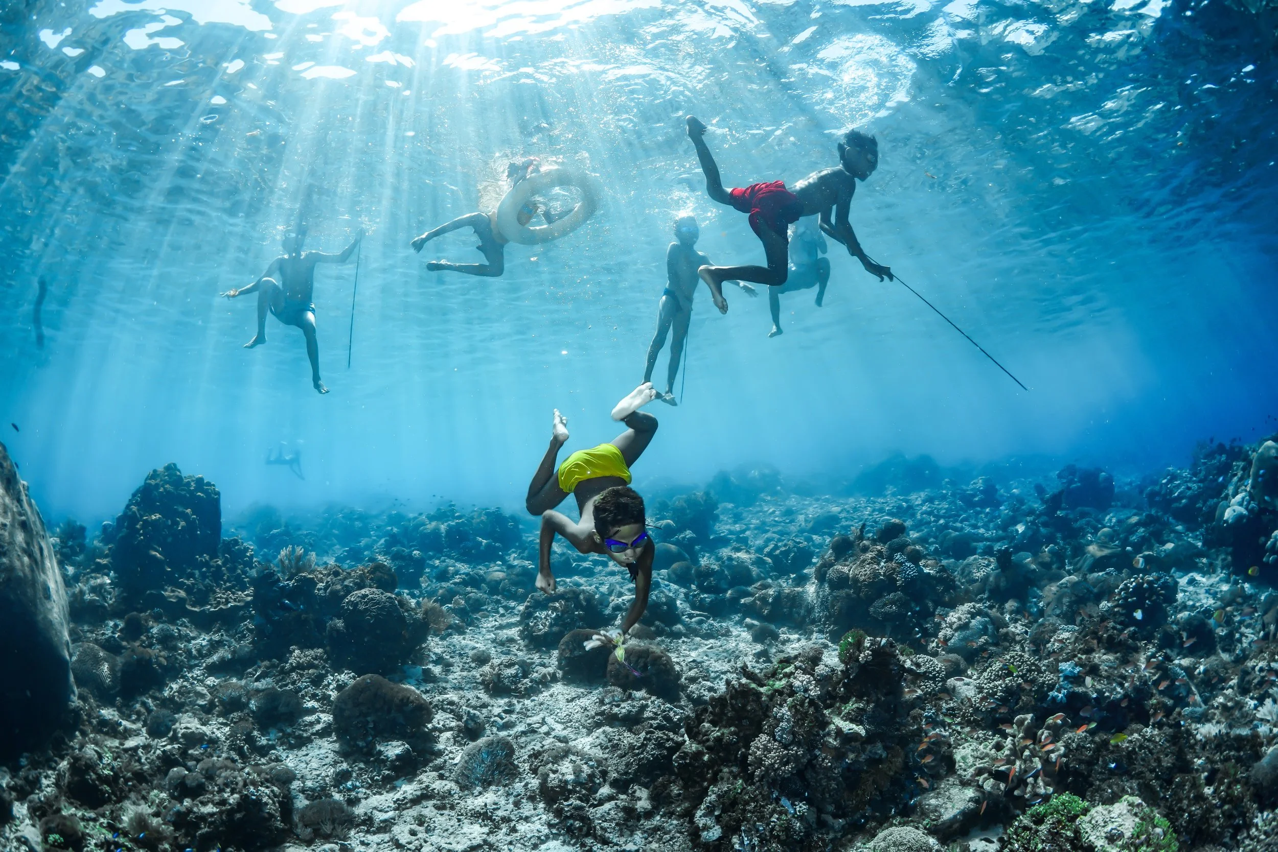 Children and teenagers swimming and playing in a clear blue ocean above a coral reef, sunlight streaming through the water.