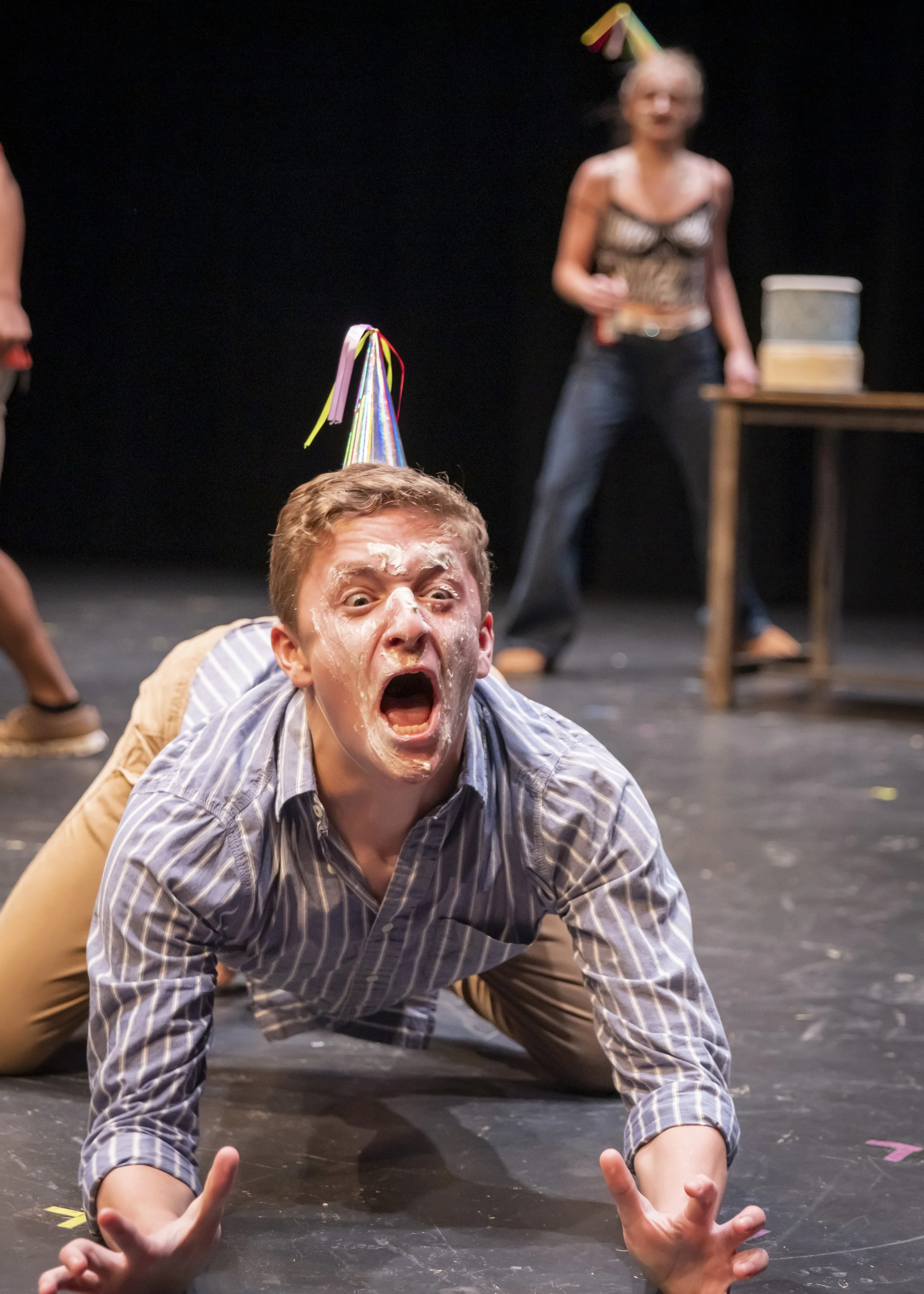 A man with a party hat, face messy with whipped cream, is crawling on the stage with his mouth open in a dramatic expression. In the background, two women are standing, one wearing a leopard print top and the other partially visible, with a table nea