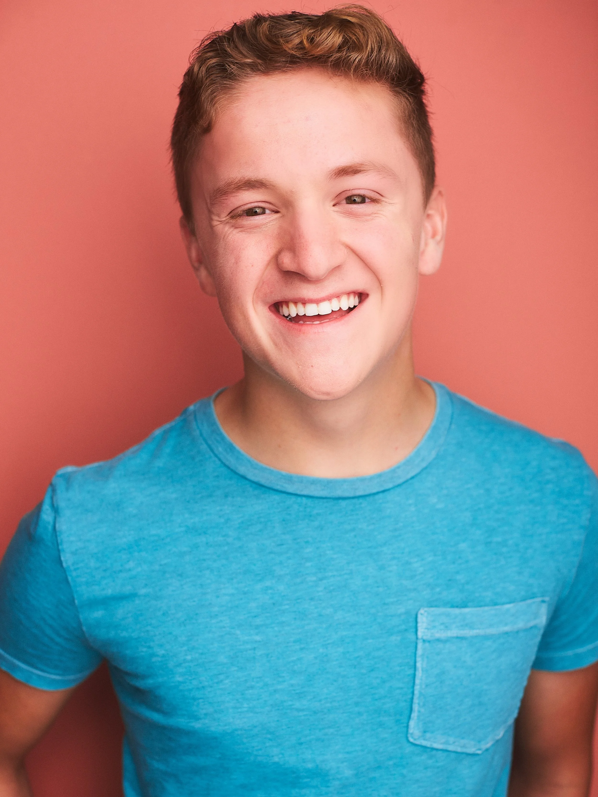 Smiling young man with short light brown hair, wearing a bright blue t-shirt, standing against a warm coral background.
