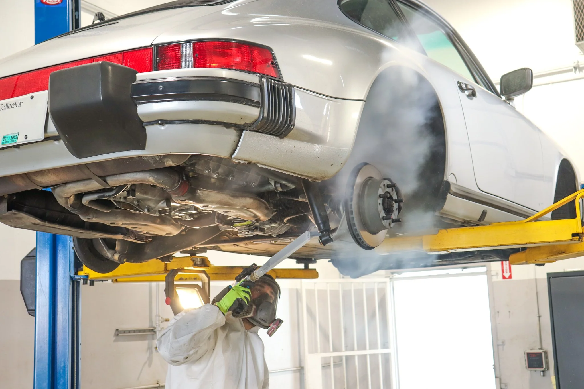 A mechanic in white coveralls and green gloves working underneath a lifted vintage silver car in a garage, with exhaust fumes visible.