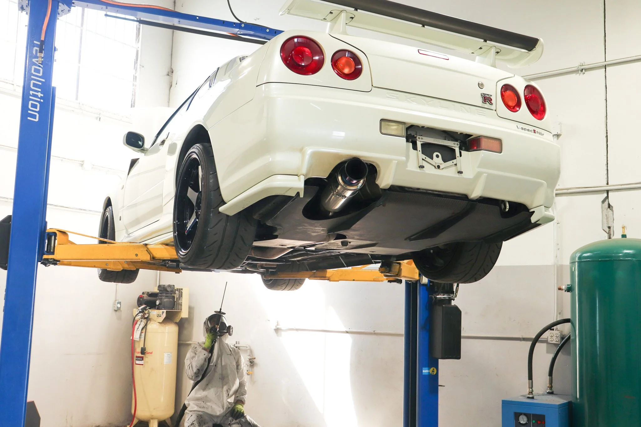 A white Nissan Skyline GT-R car is lifted on a hydraulic car lift inside a garage or workshop. A person wearing a white jumpsuit and gloves is working underneath the car. The workspace has various equipment including a large tank, a green pressure vessel, and tools.