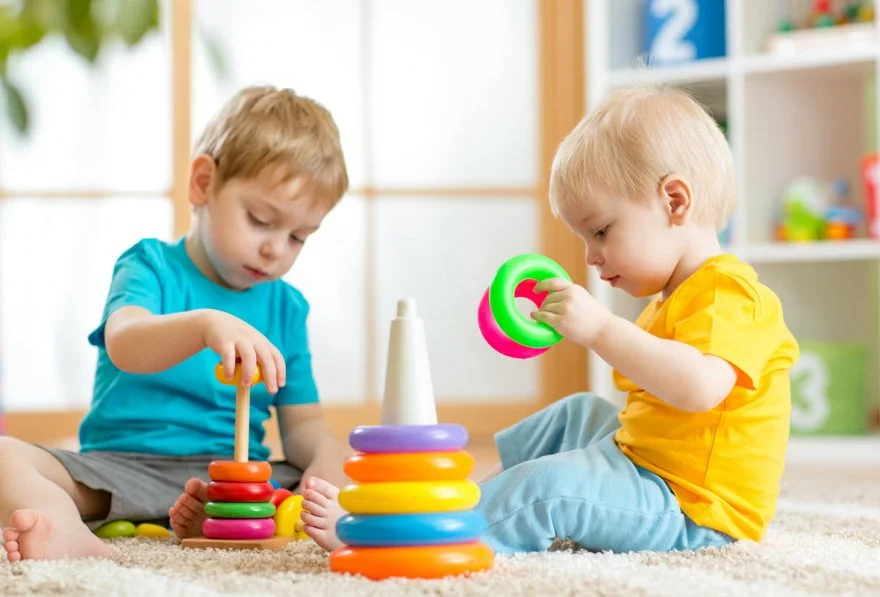Two young boys playing with colorful stacking rings on a carpeted floor in a playroom.