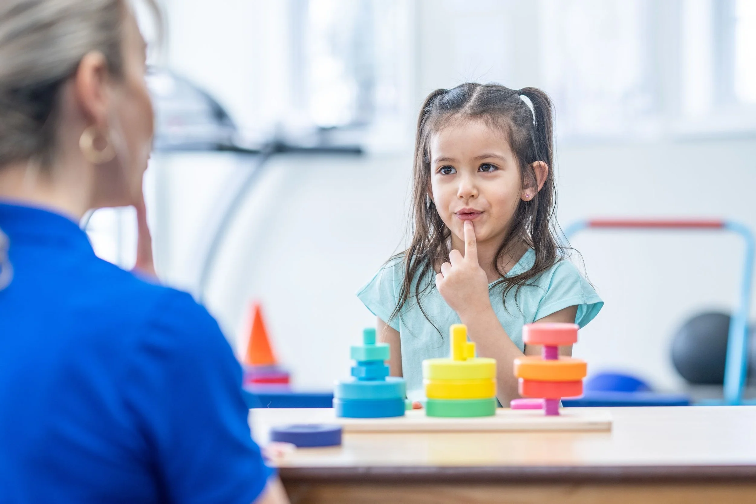 A young girl with long brown hair and pigtails, sitting at a table with colorful stacking toys, thinking with her index finger on her lips during a therapy session with a therapist whose back is to the camera.