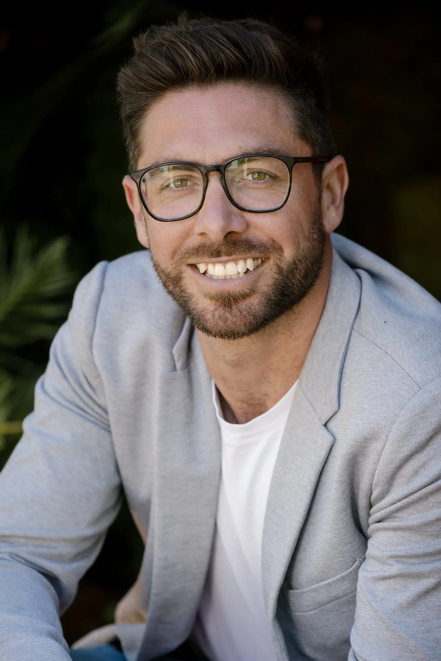 Portrait of smiling man with glasses, beard, and short hair, wearing a light gray blazer and white shirt, outdoors with green foliage in background.