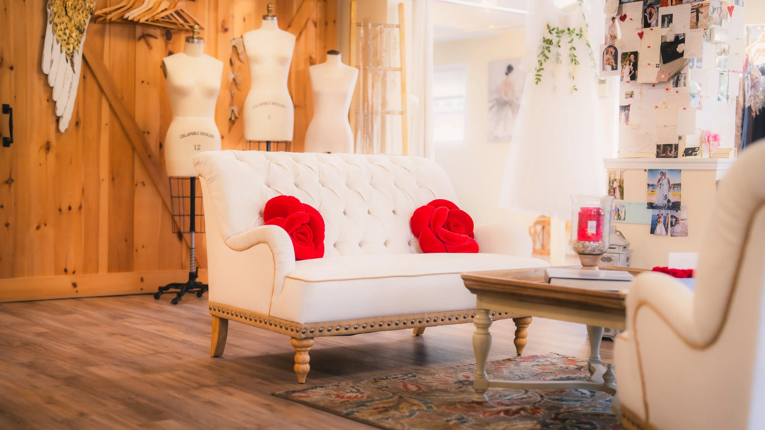 A cozy indoor space with a white tufted sofa decorated with two large red rose-shaped cushions, a light wood coffee table with a red candle and books, a white armchair, a wall with photos and notes, three mannequin dress forms, and a mannequin dressed in a white dress in the background, all set against wood-paneled and white walls with natural light.