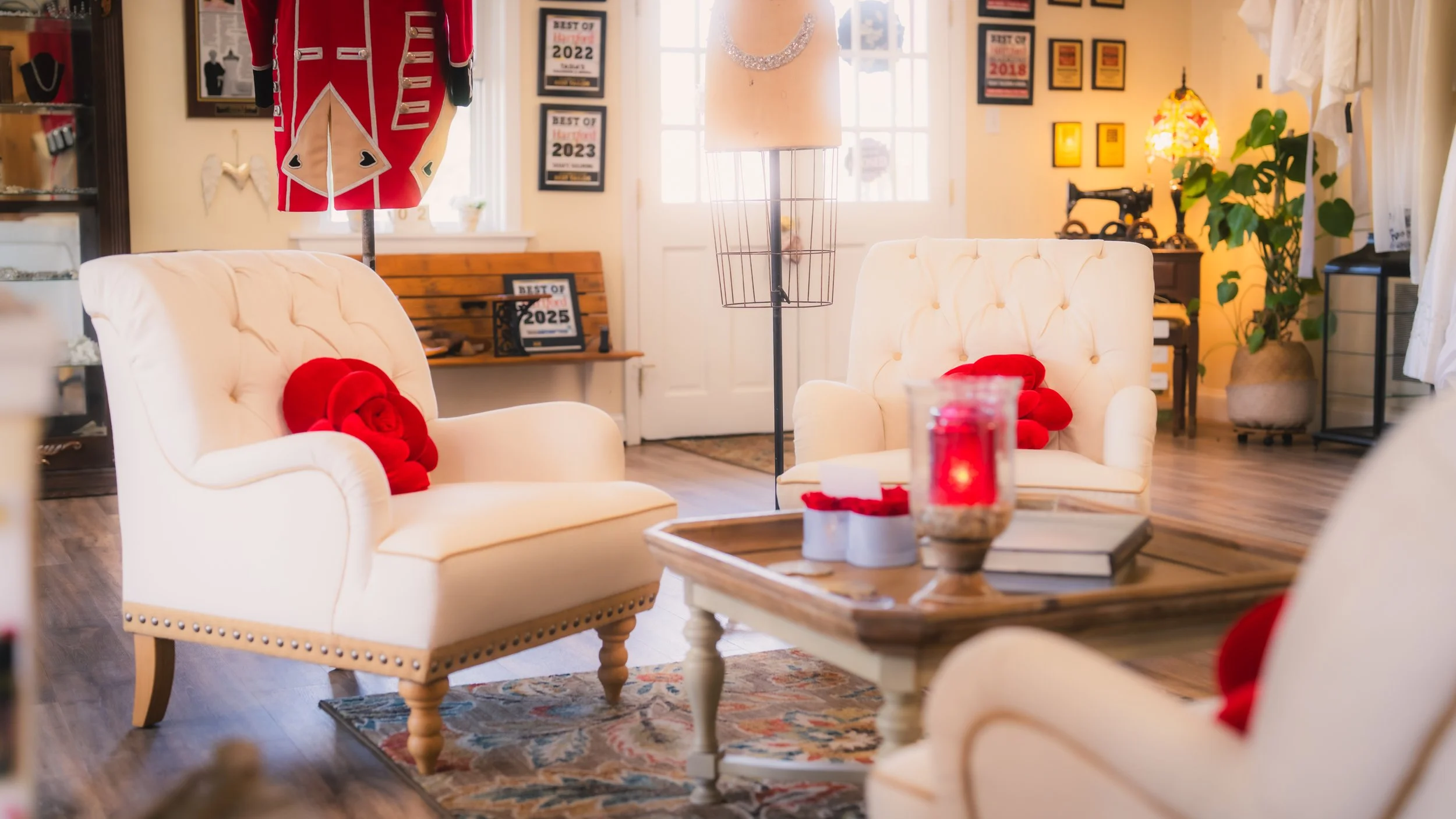 Cozy sitting room with white armchairs, red heart-shaped pillows, a wooden coffee table with candles and books, behind is a wall with framed awards, a mannequin with a silver collar, a tall lamp, and a large potted plant.